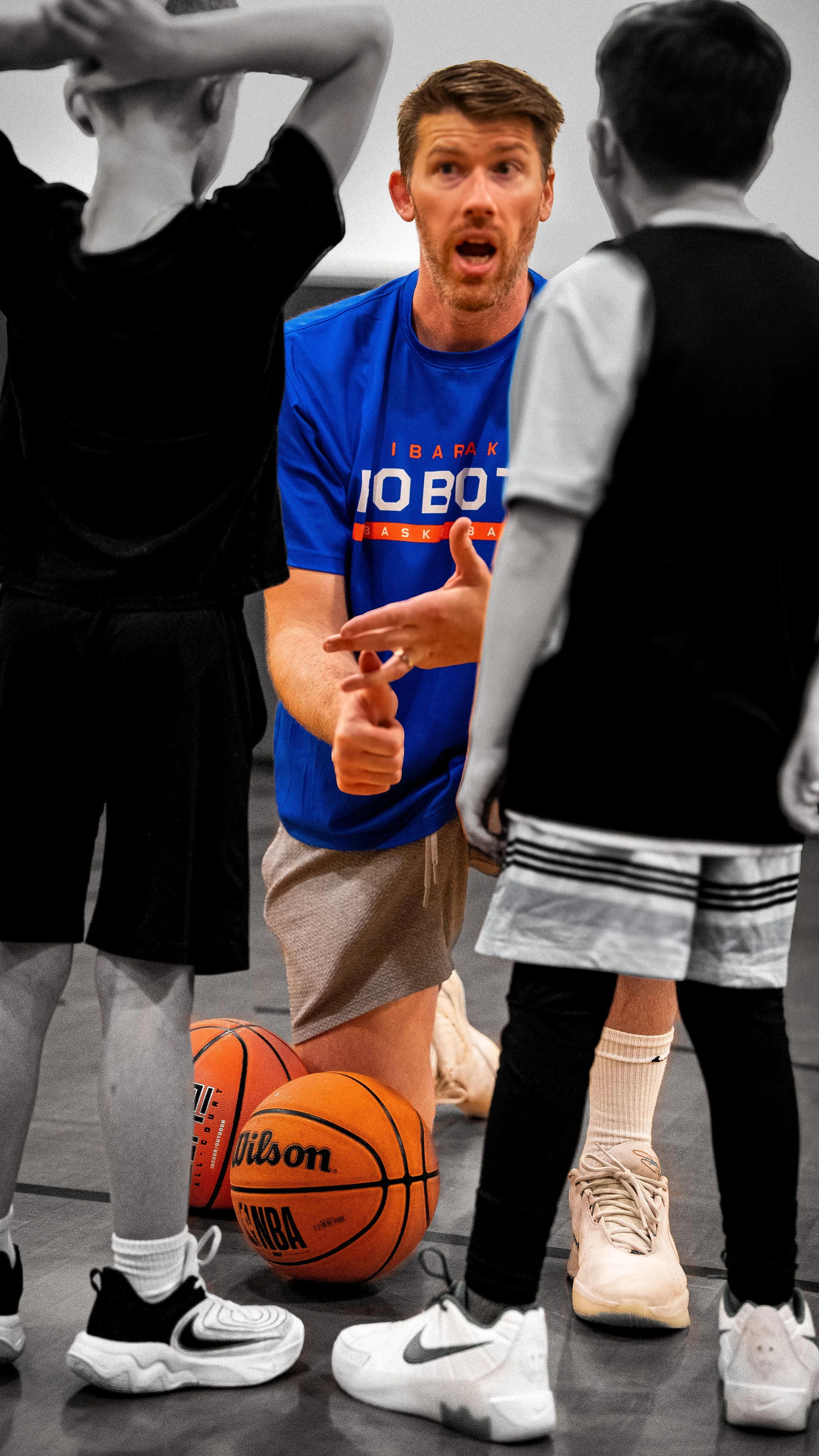 A basketball coach speaking to players during practice, with basketballs on the floor and players in athletic shoes standing in a gym.