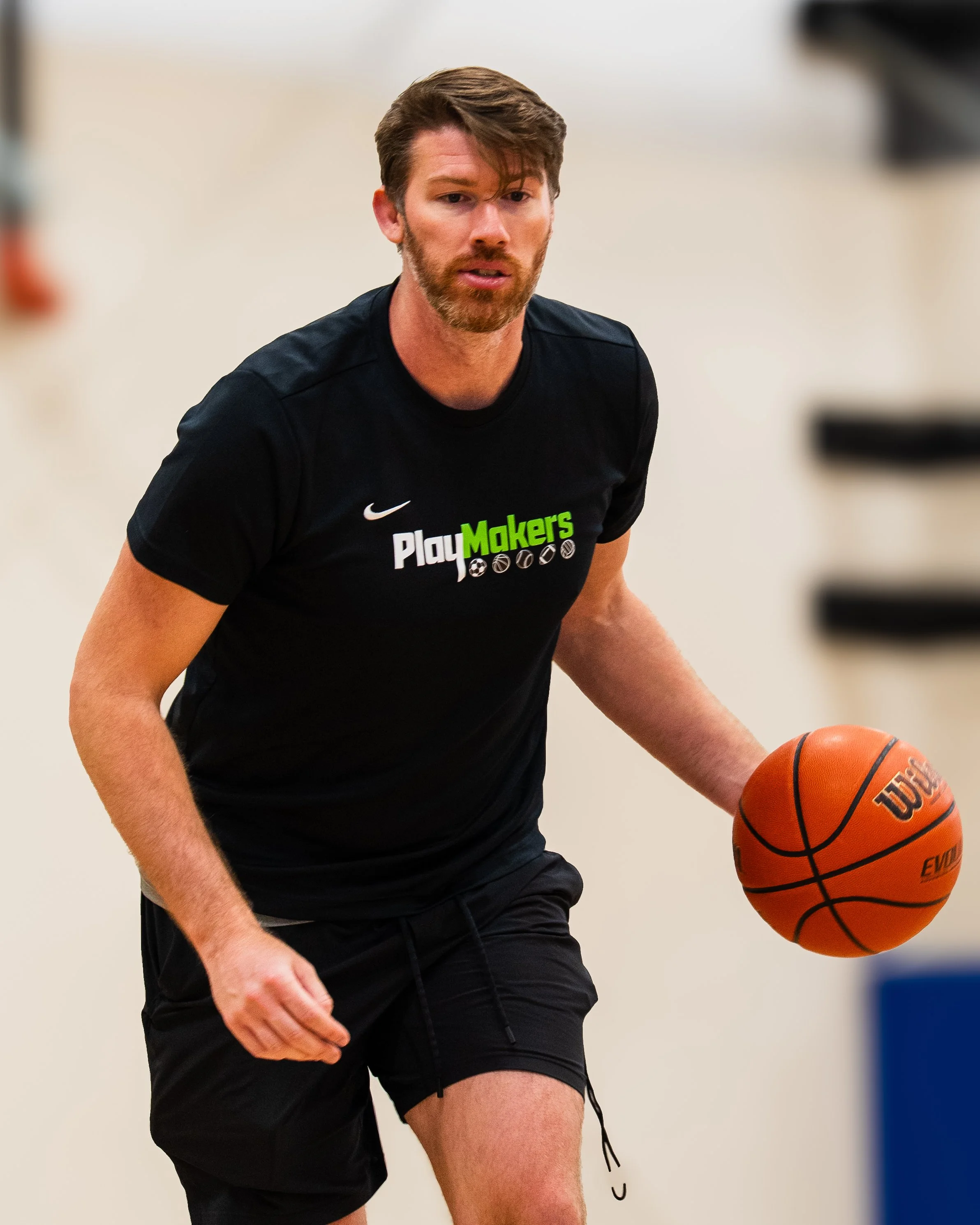A man playing basketball indoors, wearing a black shirt with 'PlayMakers' logo, holding a basketball.