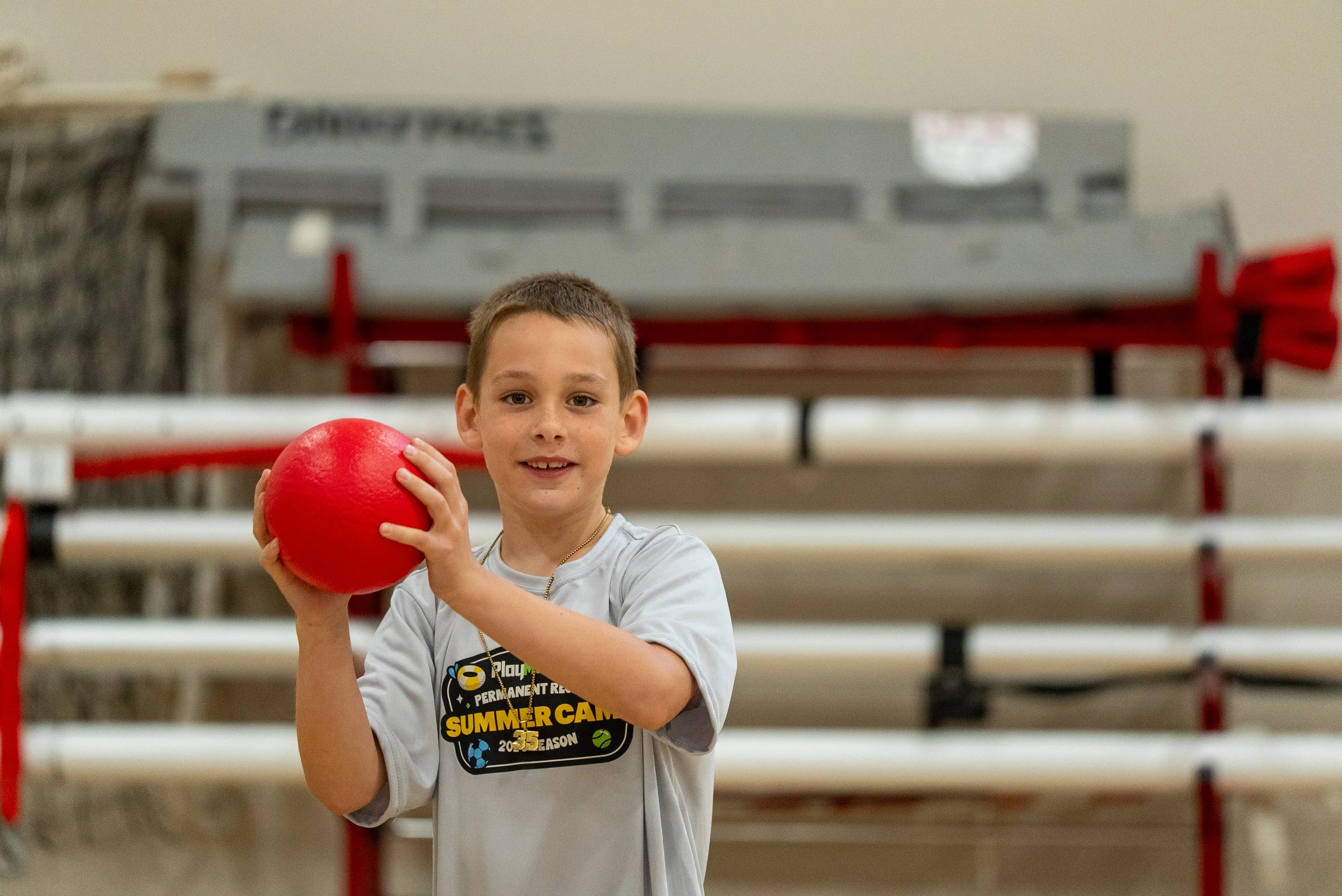 A young boy smiling and holding a red ball in an indoor gym or sports facility with bleachers in the background.