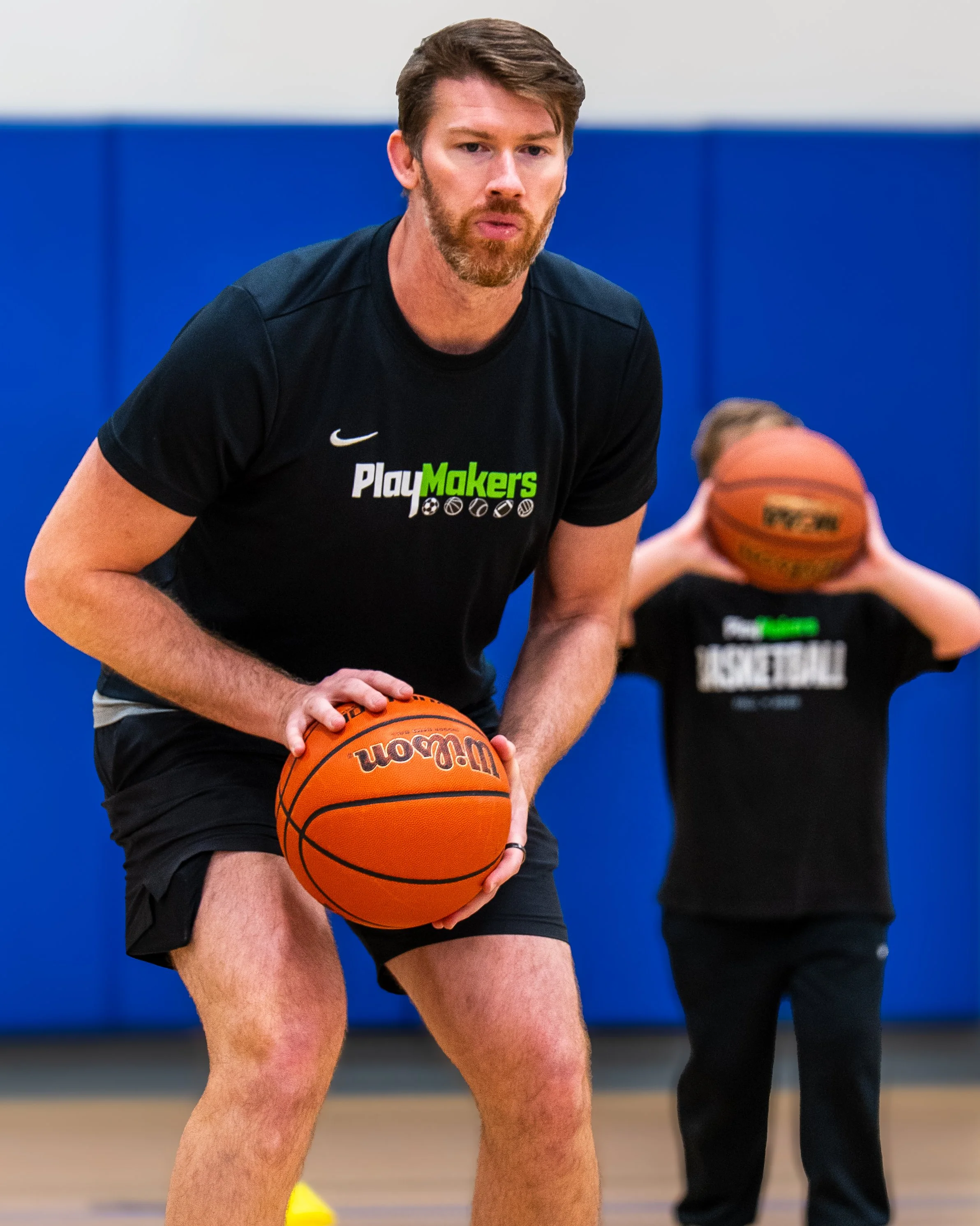 Young man with a beard holds a basketball during a practice or game in an indoor gym, wearing a black shirt with green and white text.