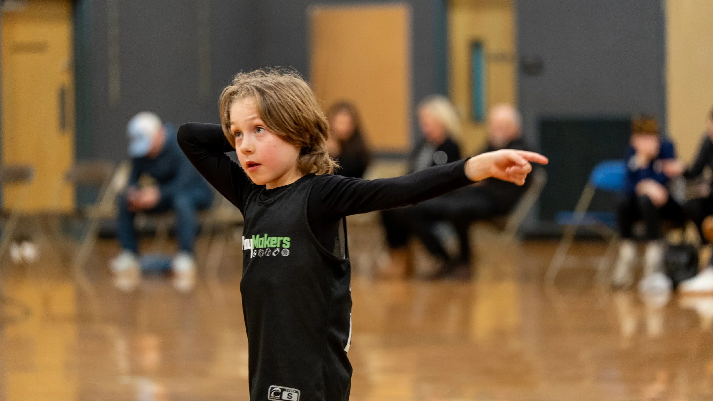 Young boy with brown hair and blue eyes standing in a gymnasium or dance studio, wearing a black long-sleeve shirt, with other people blurred in the background sitting on chairs.
