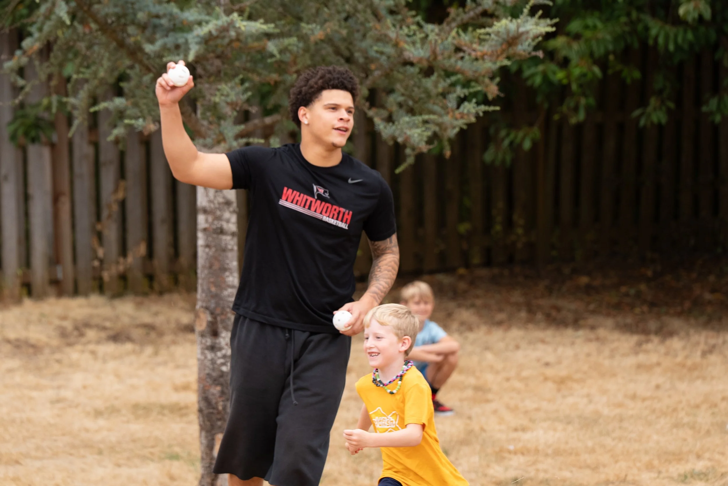 A young man with curly hair holding a white ball in each hand, standing outdoors with children around him. He is wearing a black t-shirt with red and white text and black pants. The children and the man are in a playful setting with trees and a woode