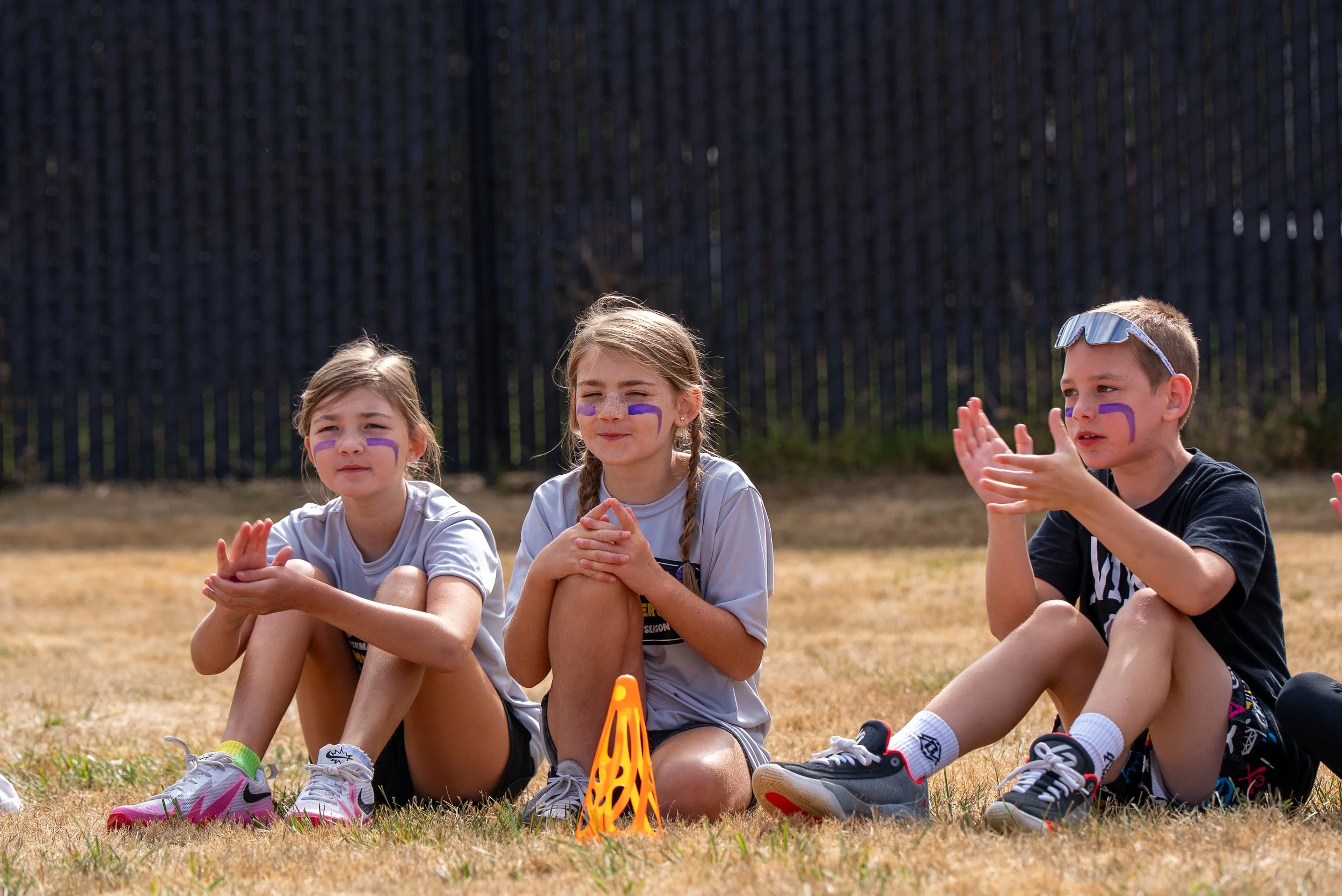 Three children sitting on the grass during an outdoor event, wearing sports attire and face paint, with orange relay batons in front of them.