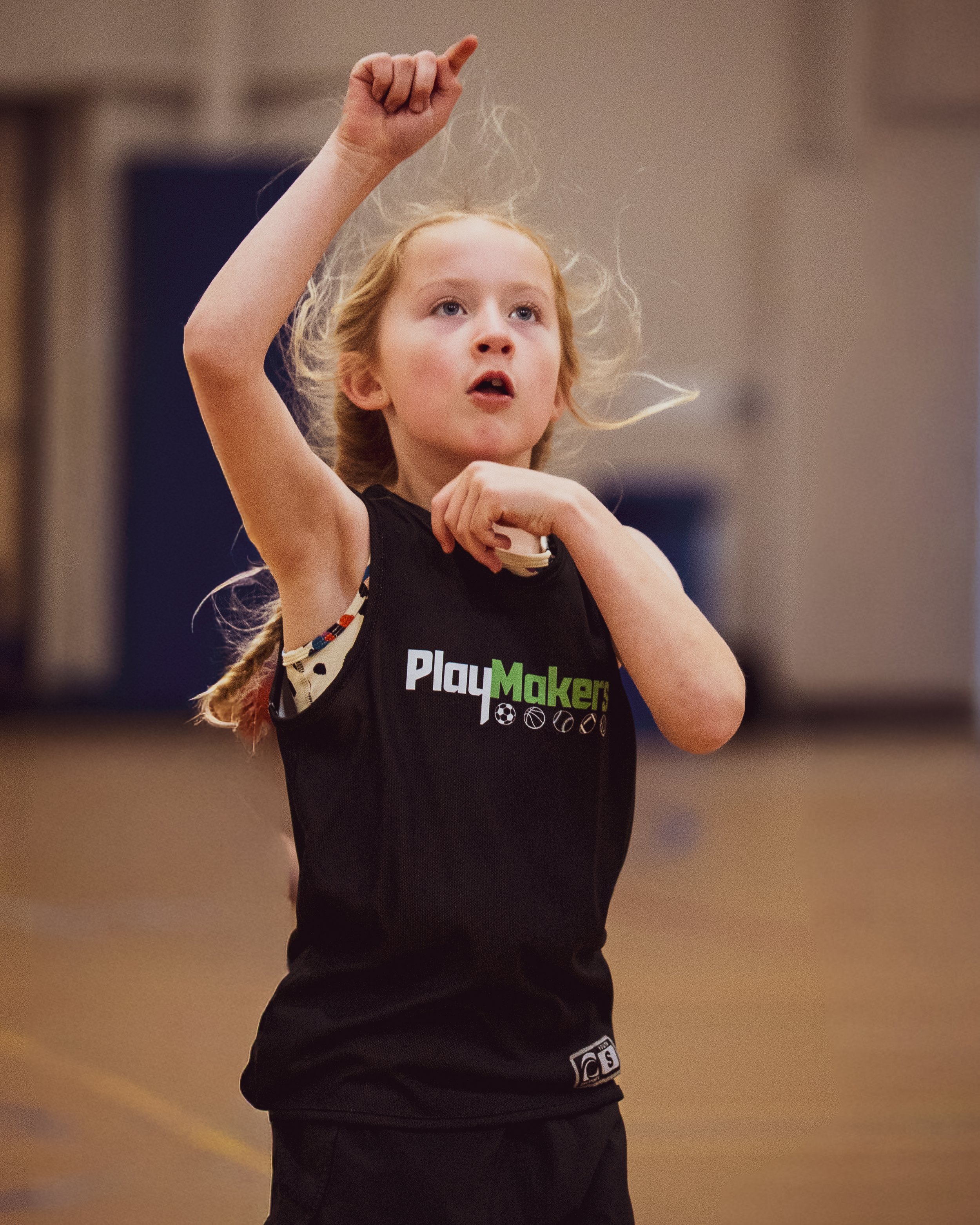 Young girl with blonde hair and fair skin in a black sports jersey with the logo 'Play Maker', raising her right arm with one finger pointing upwards during an indoor sports activity.