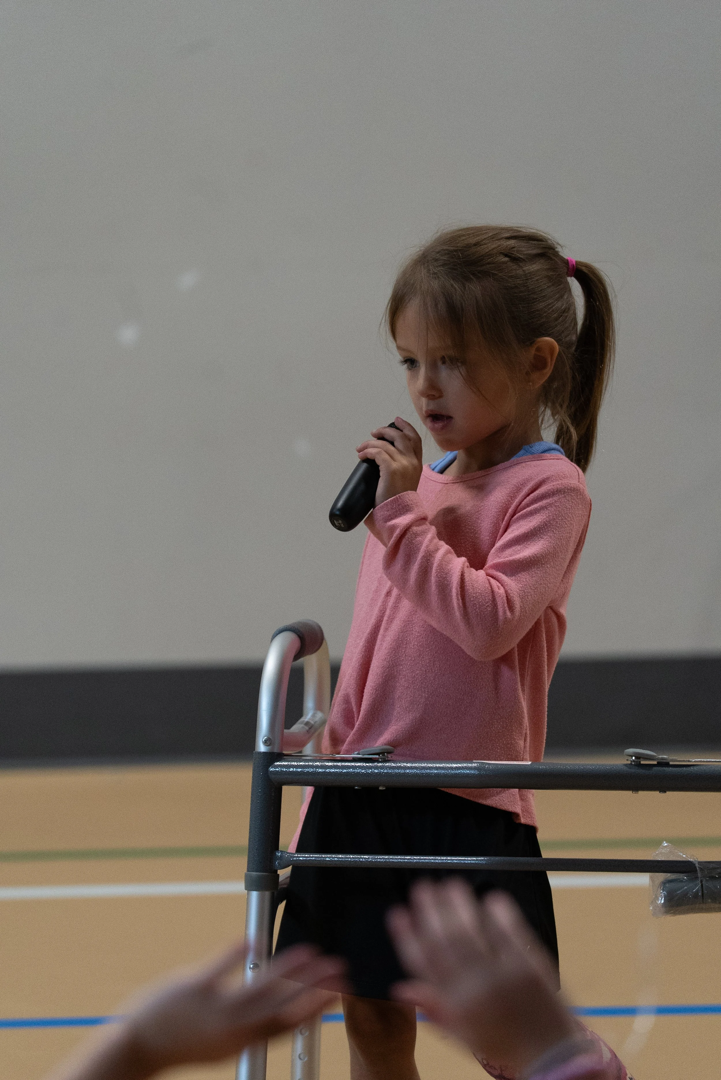 A young girl with brown hair tied in a ponytail, wearing a pink long-sleeve shirt, holding a microphone and standing behind a walker.