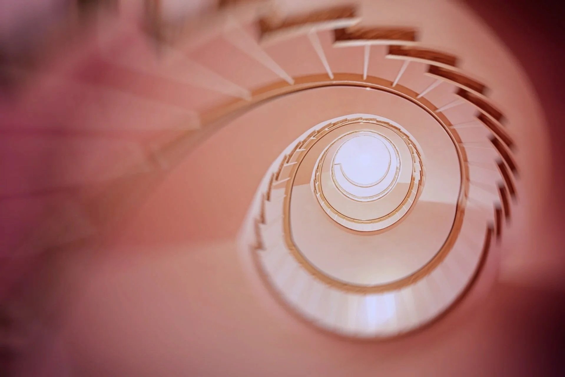 A view looking down a spiral staircase with orange-colored steps and railings, creating a tunnel-like effect toward a bright light at the bottom.