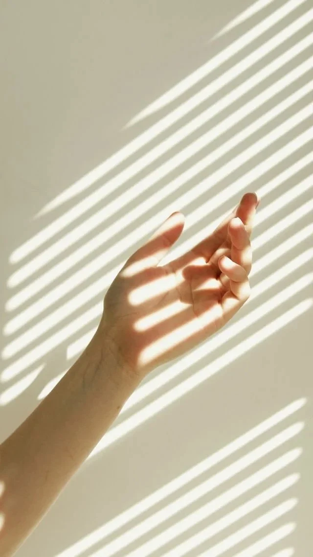 A human hand with fingers slightly bent, casting striped shadows from sunlight passing through window blinds onto a plain wall.