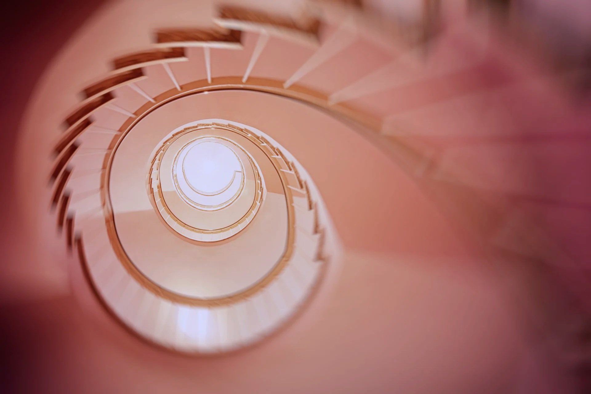 View of a spiral staircase looking down from the top, featuring a pinkish hue and wooden railings.