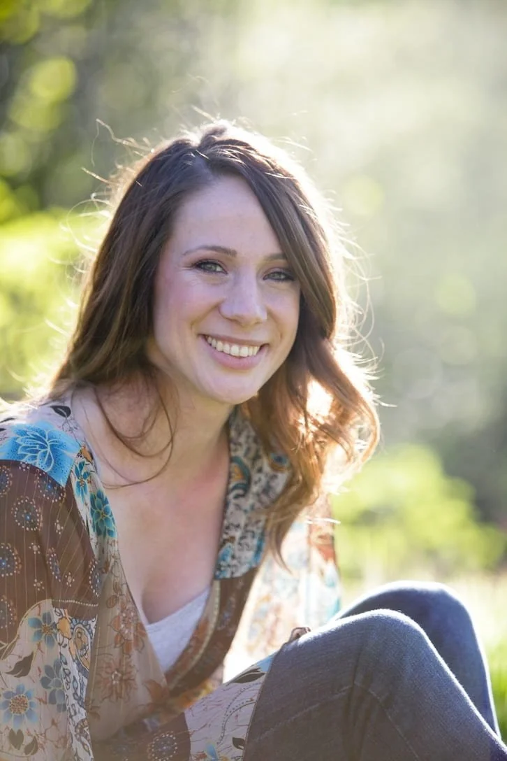 Young woman sitting outdoors in sunlight, smiling, with green foliage in the background.