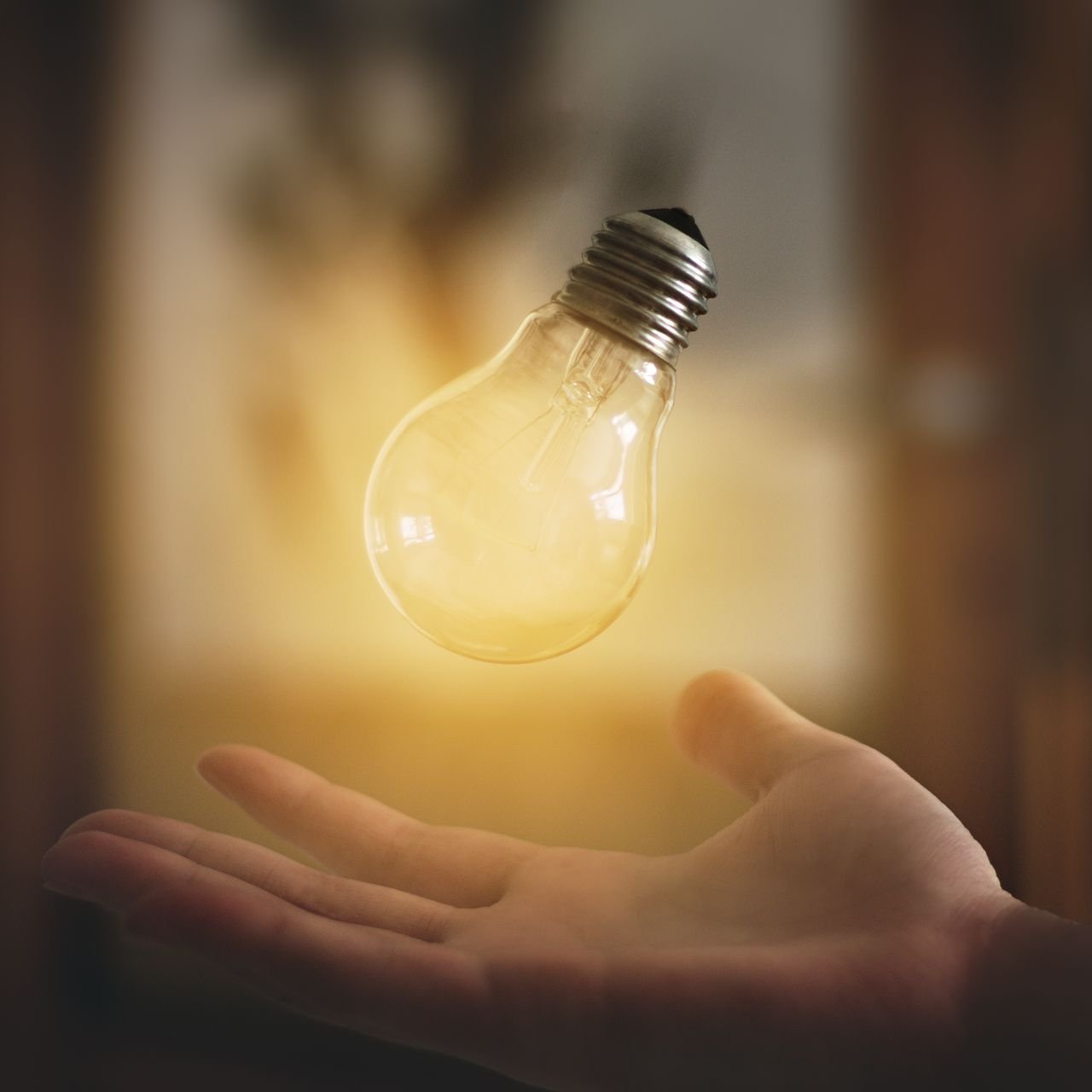A hand holding a floating light bulb with a glowing filament in a blurred indoor background.