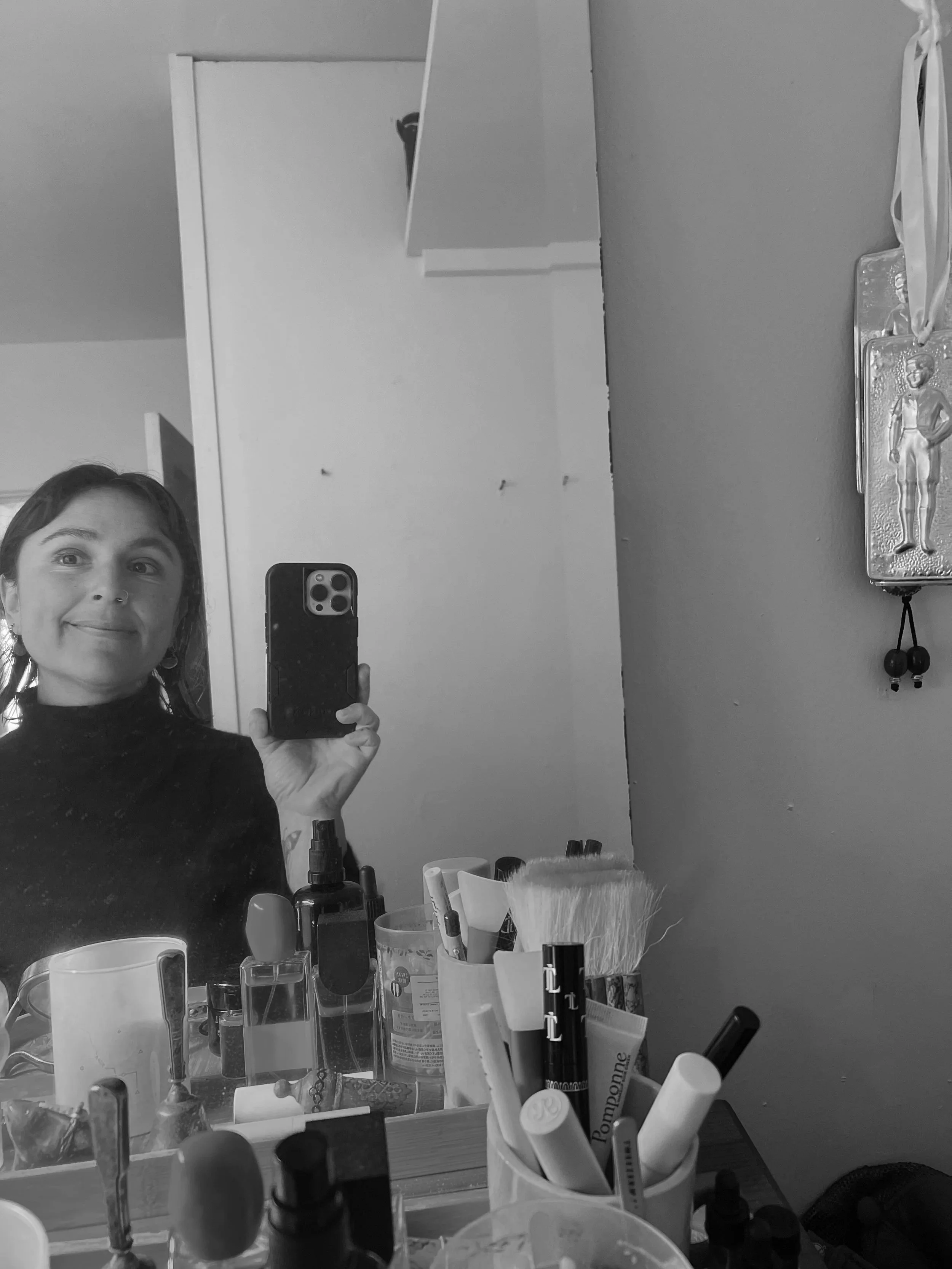 A woman taking a selfie in her bathroom mirror, with various toiletries and personal care items on the bathroom counter. The mirror reflects part of her face and the bathroom background.