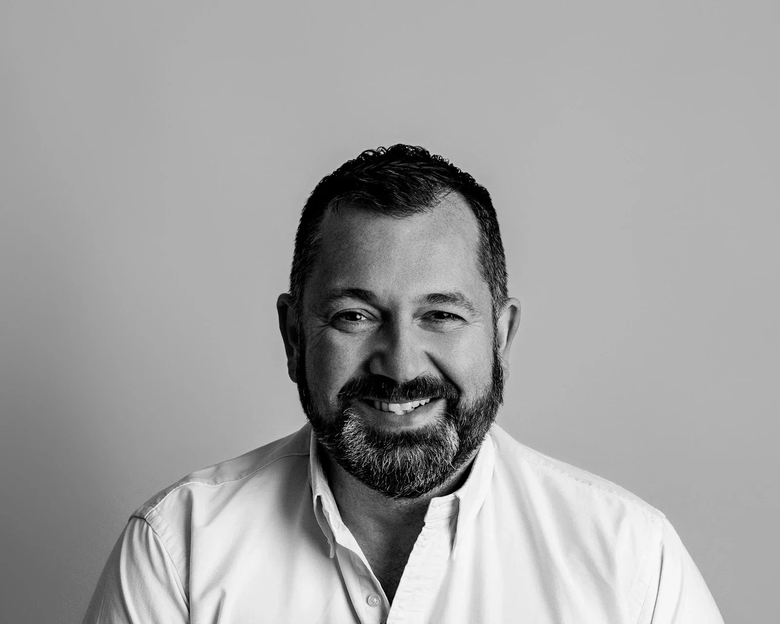 Black and white portrait of a smiling man with a beard, wearing a white button-up shirt, against a plain gray background.