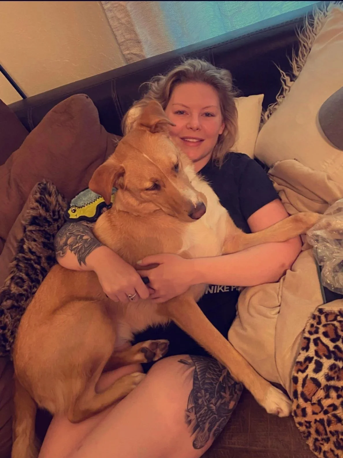 A woman lying on a couch, hugging her dog biscuit while smiling at the camera.