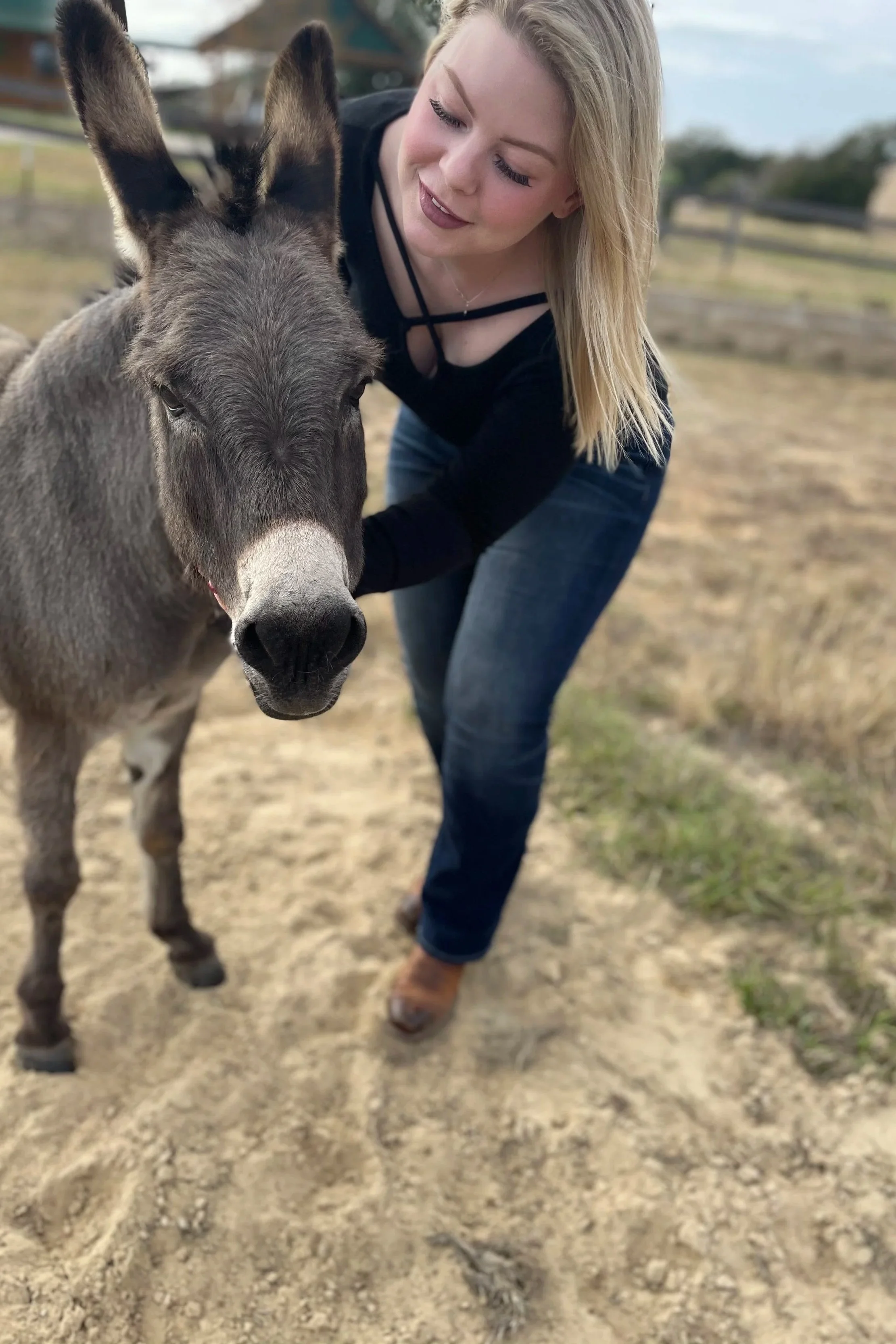 A woman with blonde hair smiling and leaning towards a donkey in an outdoor rural setting with a dirt path and fencing.