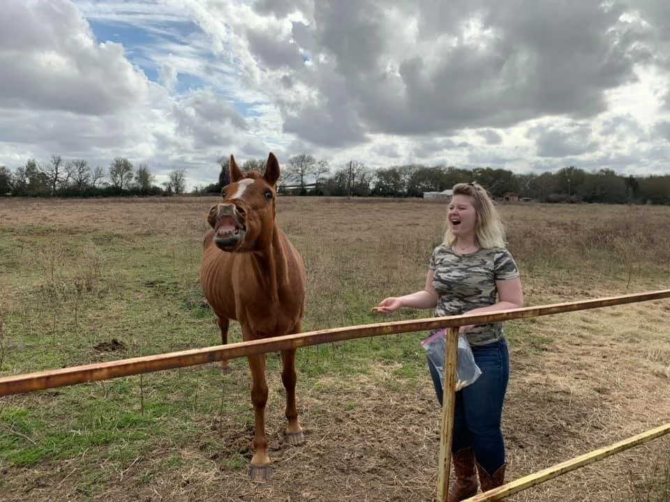 A woman laughing at a farm while standing near her horse Blaze beside a wooden fence.