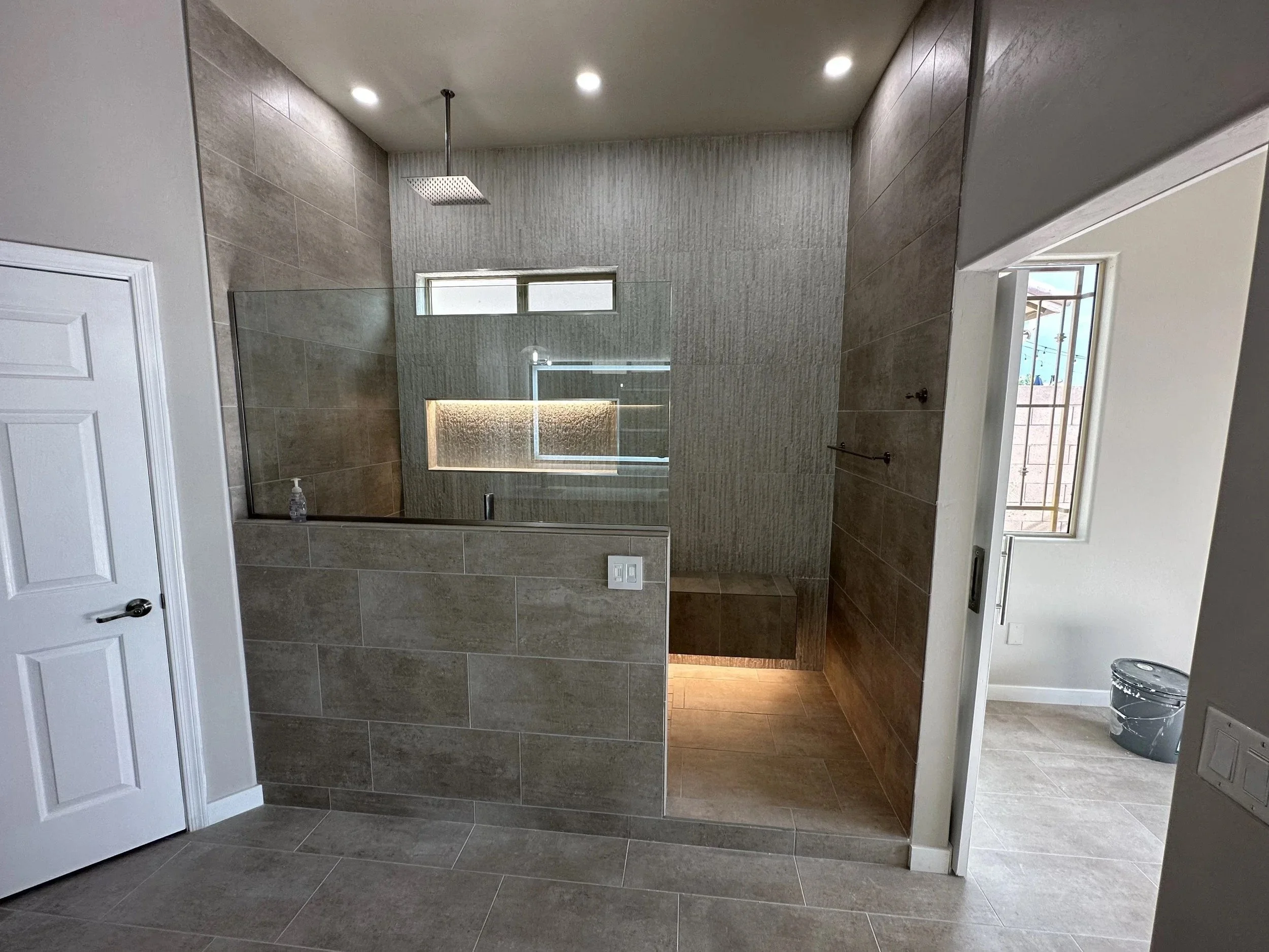 Modern bathroom featuring a walk-in shower with a glass partition, a rainfall showerhead, gray textured tiles, a window, and a white door.