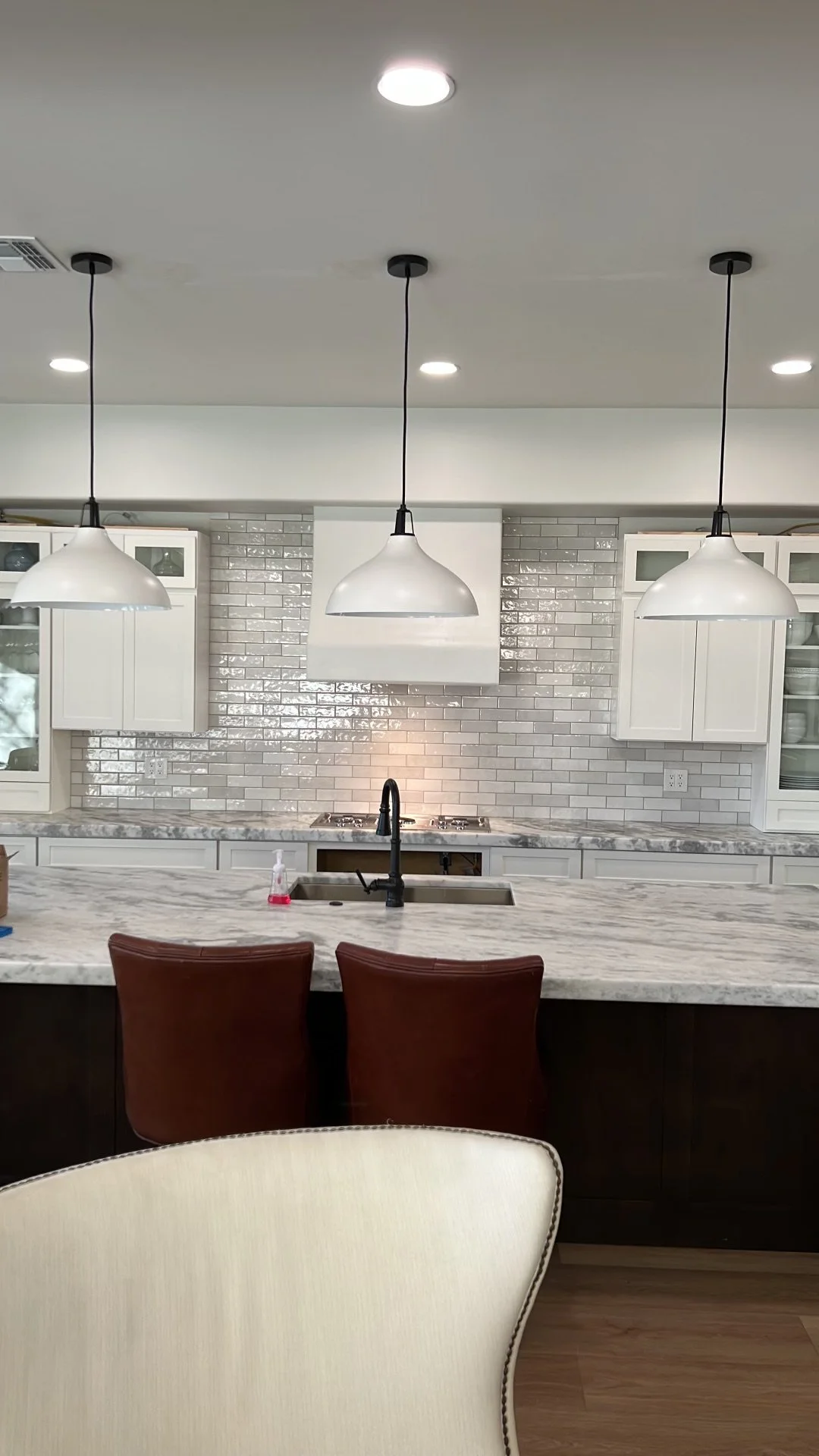 Modern kitchen with white cabinets, gray subway tile backsplash, marble countertop, pendant lights, black faucet, and brown chairs.