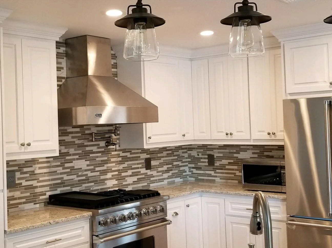 Modern kitchen with white cabinets, stainless steel range hood, gas stove, microwave, and a tiled backsplash with beige and gray tones. Pendant lights hang from the ceiling.