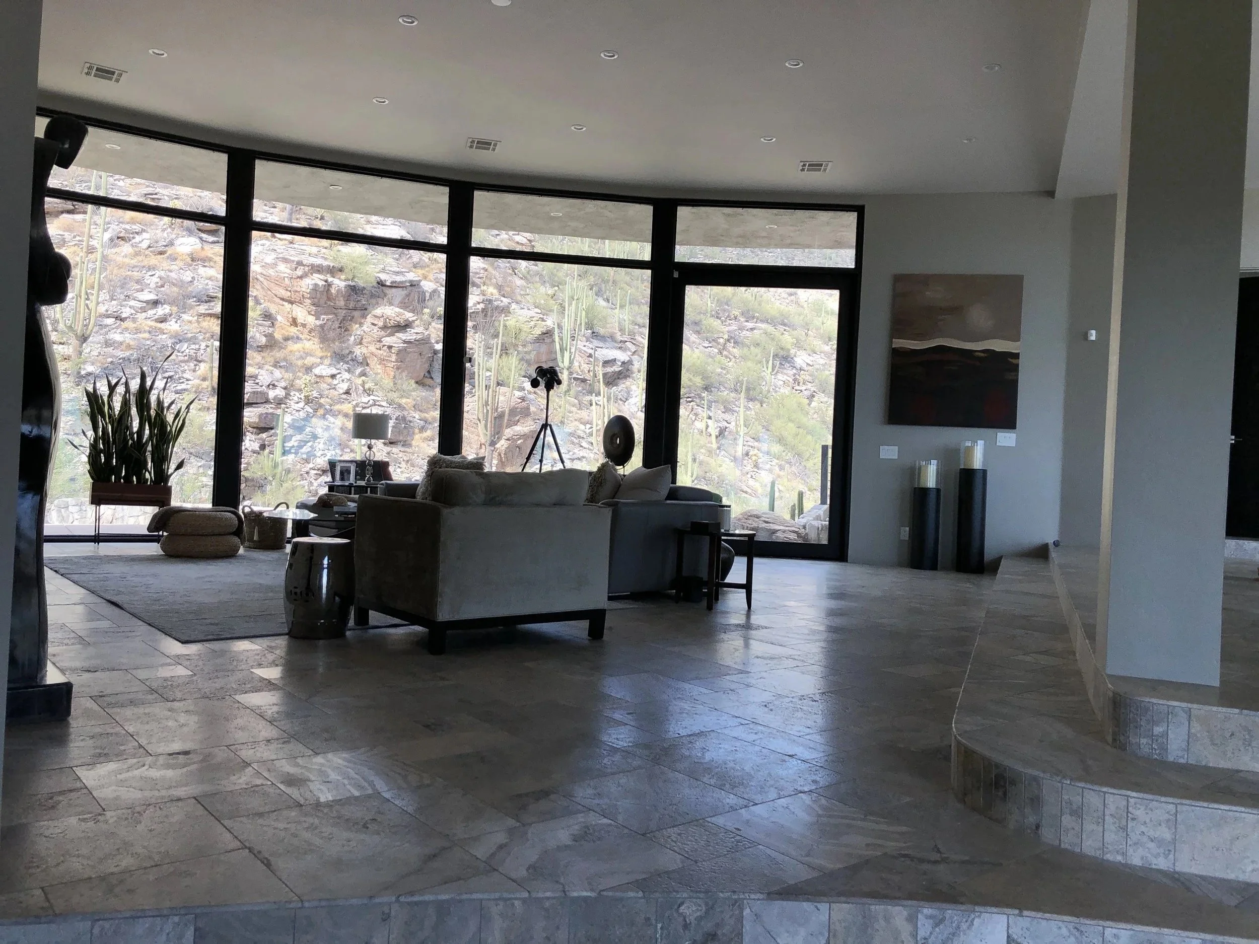 Living room with large windows overlooking a desert landscape with cacti, furnished with a white sofa, side table, tripod lamp, and a large artwork on the wall.