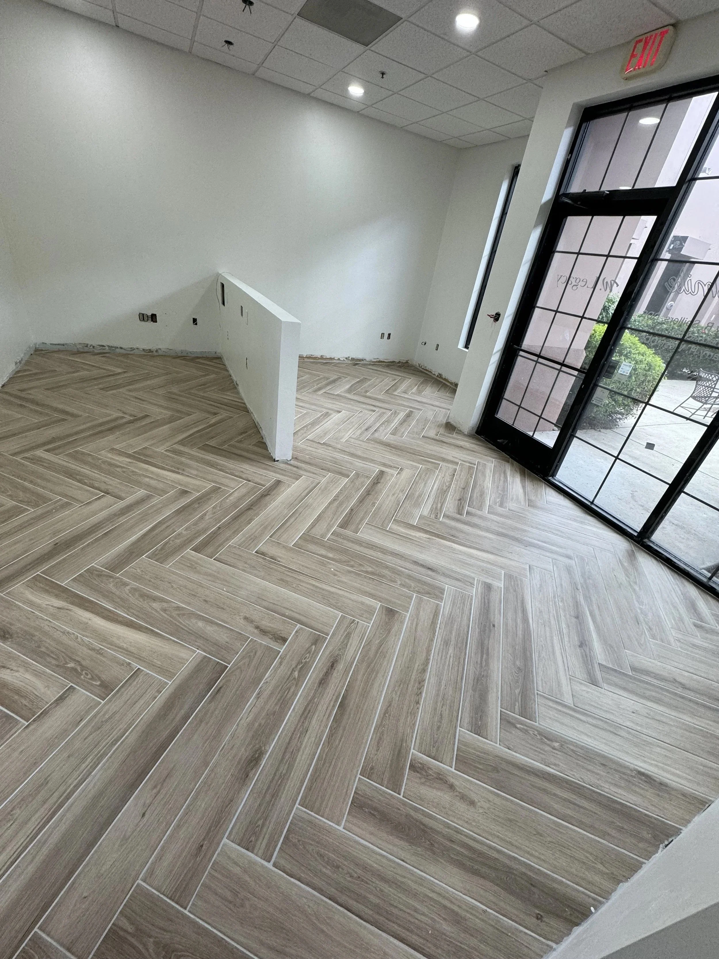 Interior space with herringbone light wood floor, white walls, ceiling with recessed lighting, and large glass door showing outside greenery.