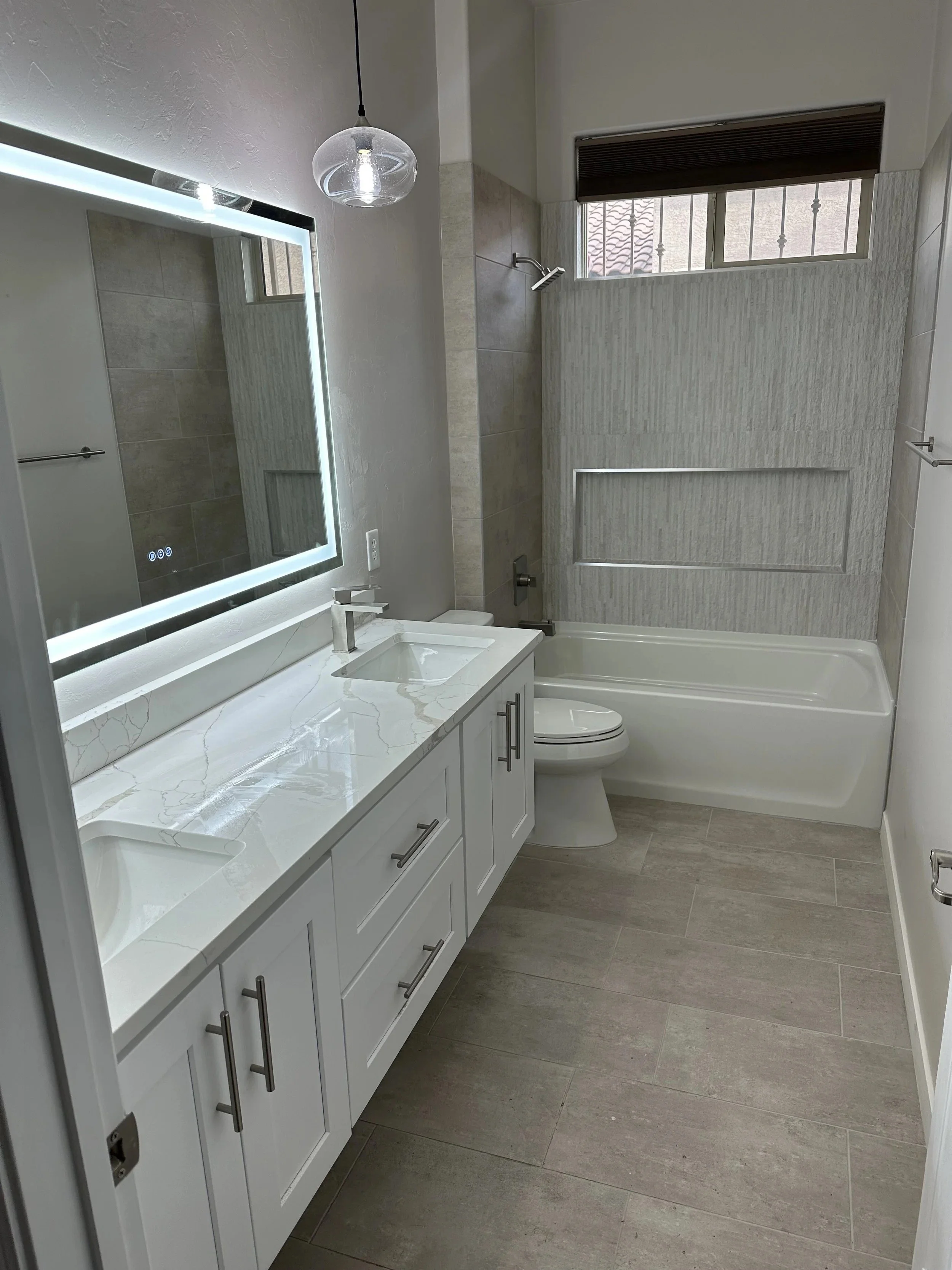 A modern bathroom with a white vanity, marble countertop, and a large illuminated mirror. A toilet is beside the vanity, and a bathtub with a shower head. A window with bars is above the bathtub, and a dark brown blind is partially drawn.