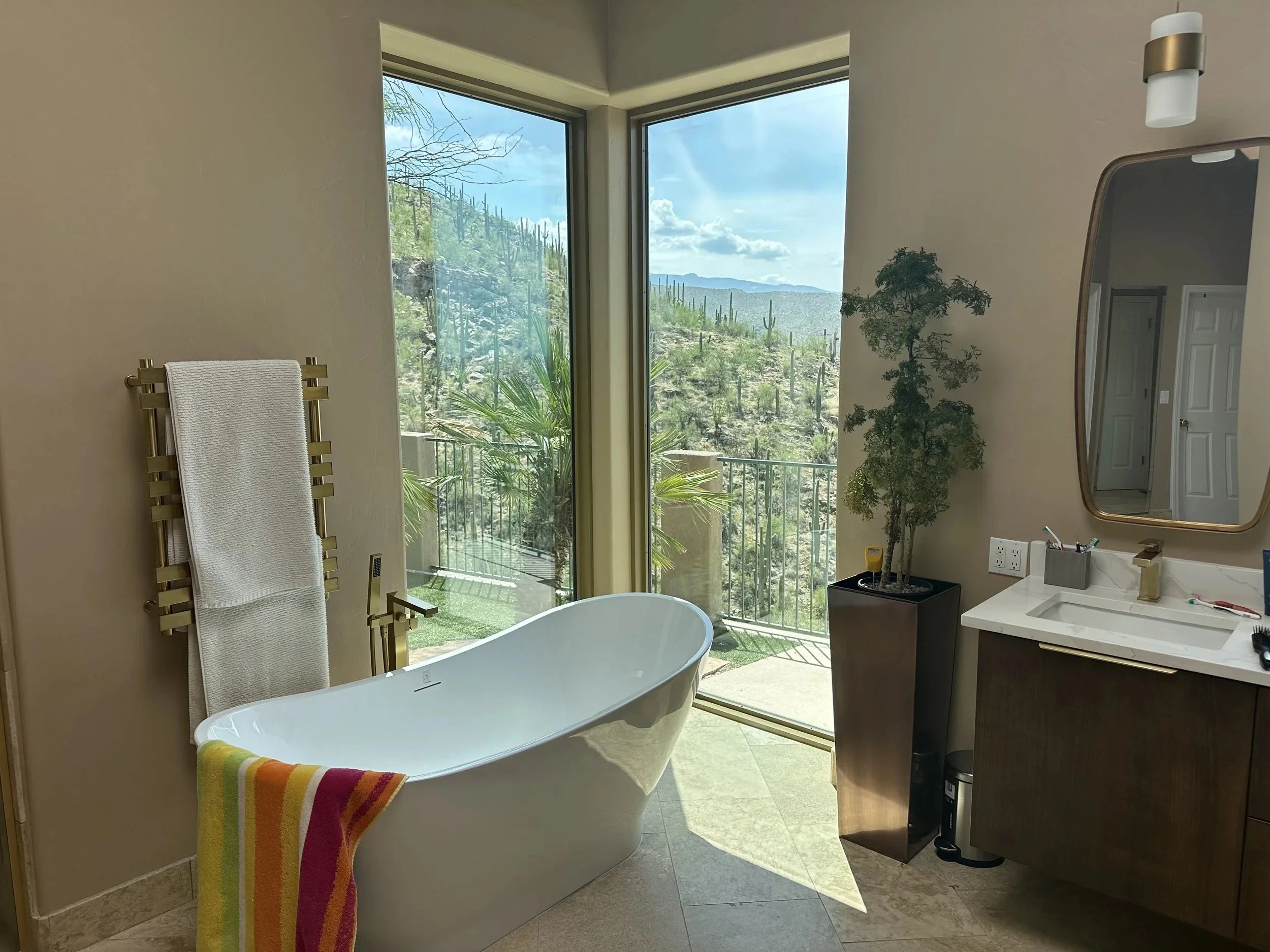 Bathroom scene with a white freestanding bathtub with a colorful towel draped over the side, large corner window showing a desert landscape with cacti and mountains, a tall potted plant on the right, a bathroom vanity with a mirror and some toiletries, and a sliding door opening to an outdoor balcony.