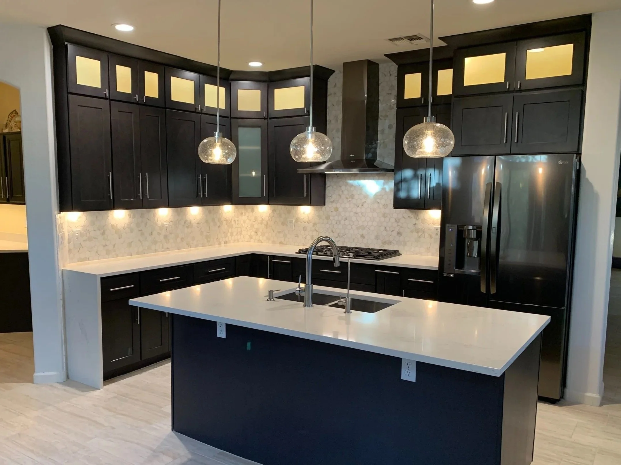 A modern kitchen with black cabinets, a white countertop island, pendant lights, and a stainless steel refrigerator.