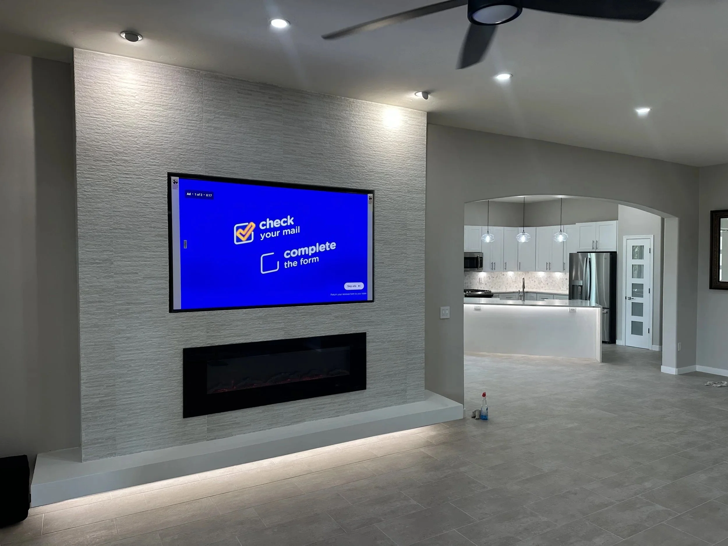 Modern living room with a wall-mounted flat-screen TV displaying a mail-checking prompt, and a built-in fireplace below the TV. The space leads into an open kitchen with white cabinetry and stainless steel appliances, illuminated by pendant lights.