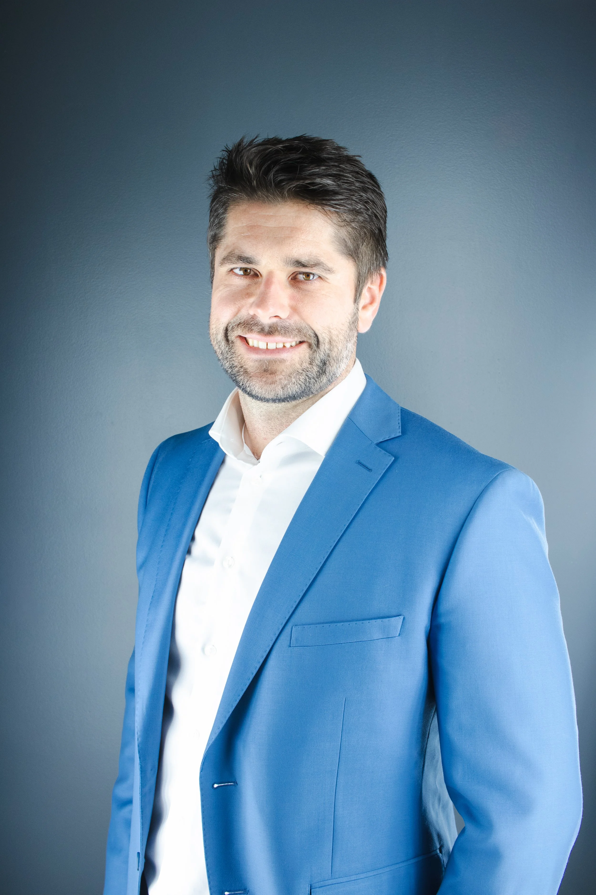 A smiling man with dark hair and a beard, wearing a white shirt and a blue blazer, standing against a gray background.