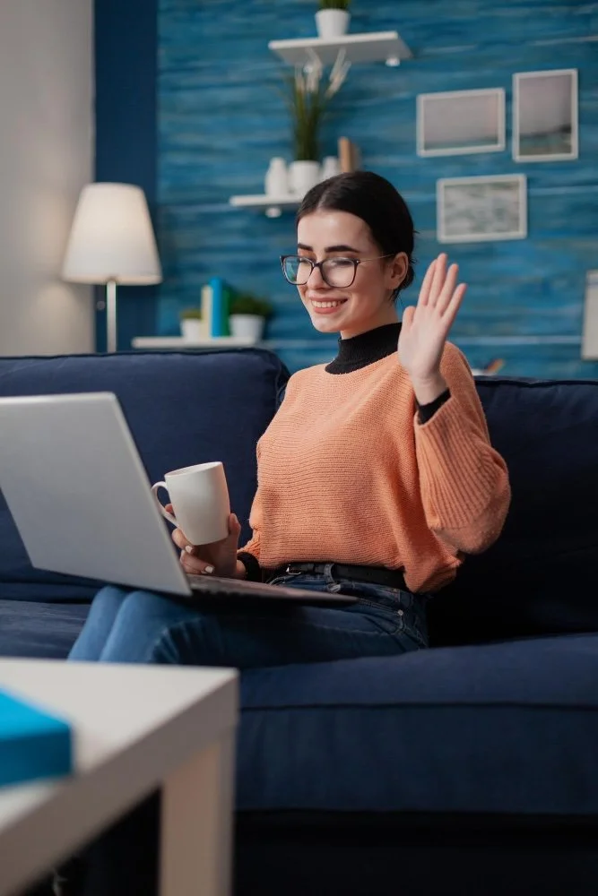 A young woman with glasses, dark hair, and a peach sweater holding a coffee mug, sitting on a blue sofa, waving at a laptop screen in a cozy, modern living room with blue walls and decorative shelves.