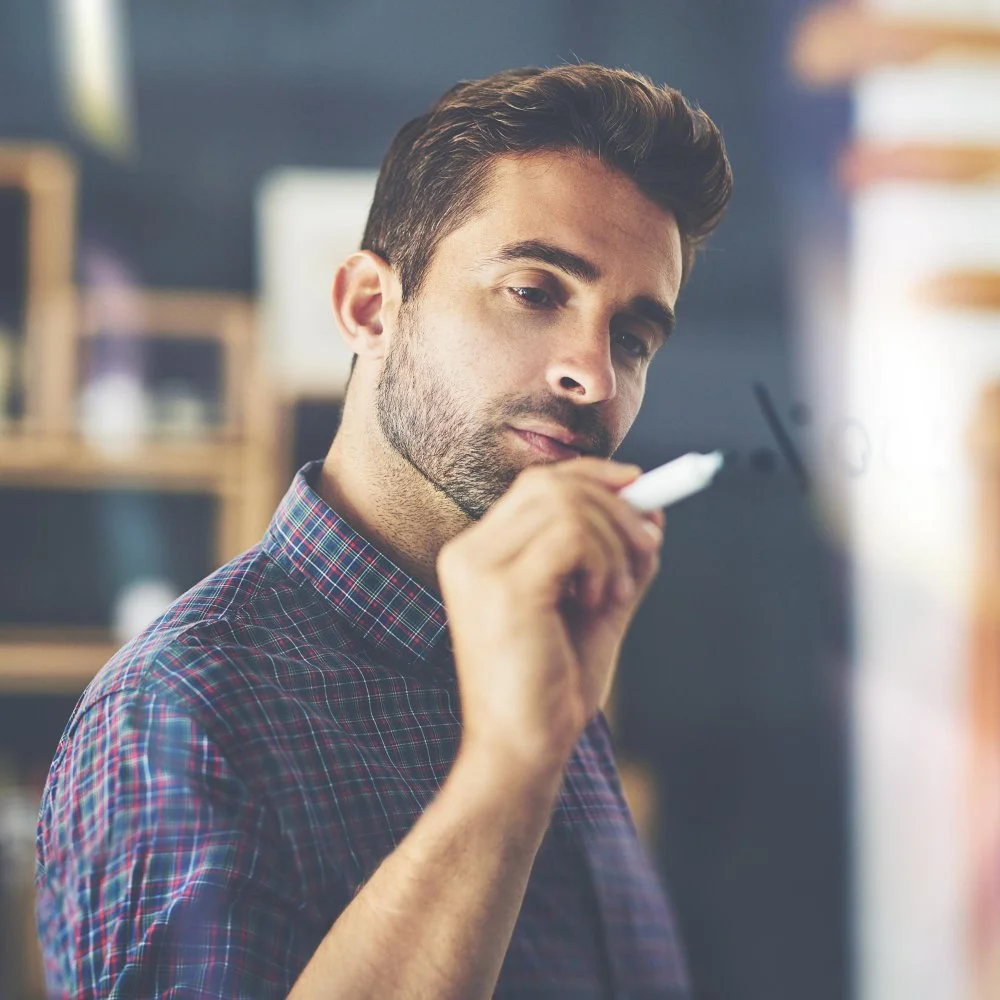 A man with dark hair and a beard wearing a plaid shirt, writing on a glass surface with a white marker.