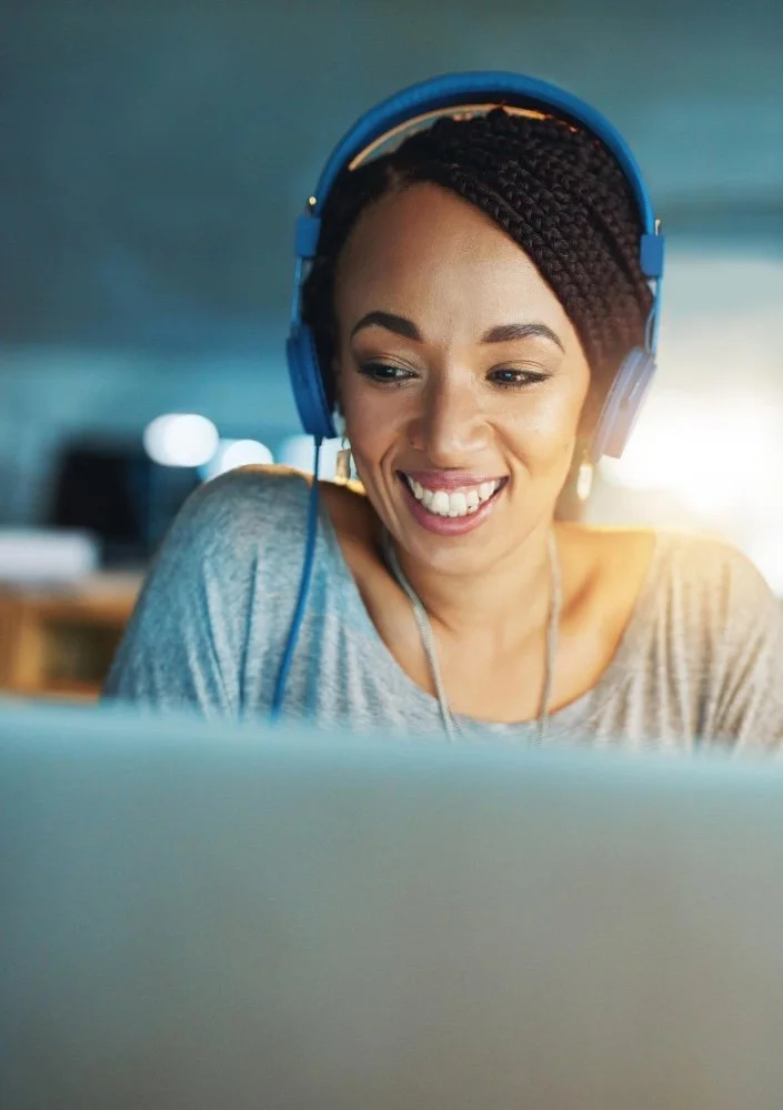 A woman with braided hair wearing blue headphones, smiling while looking at a laptop.