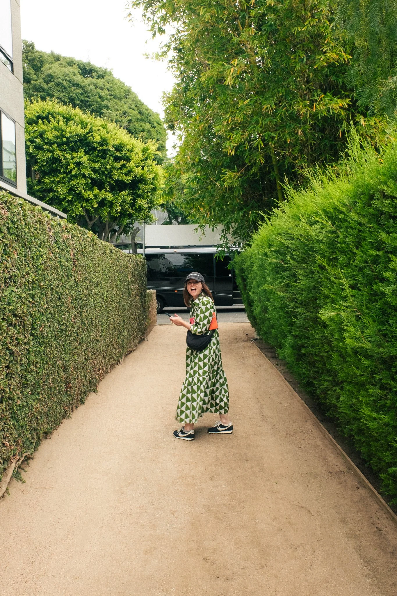 A woman with long hair, wearing a black cap, glasses, a green and white geometric patterned dress, white sneakers, and carrying a black crossbody bag, smiling and holding a phone, walking on a paved sidewalk between lush green hedges and trees.