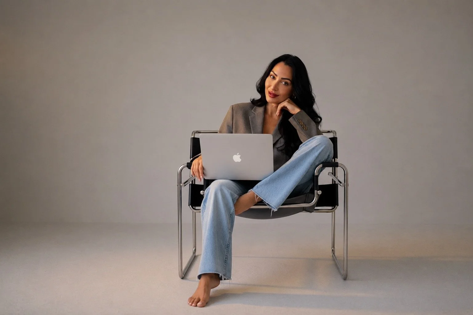 A woman sitting on a modern chair with a laptop on her lap, wearing a blazer and jeans, with a neutral background.