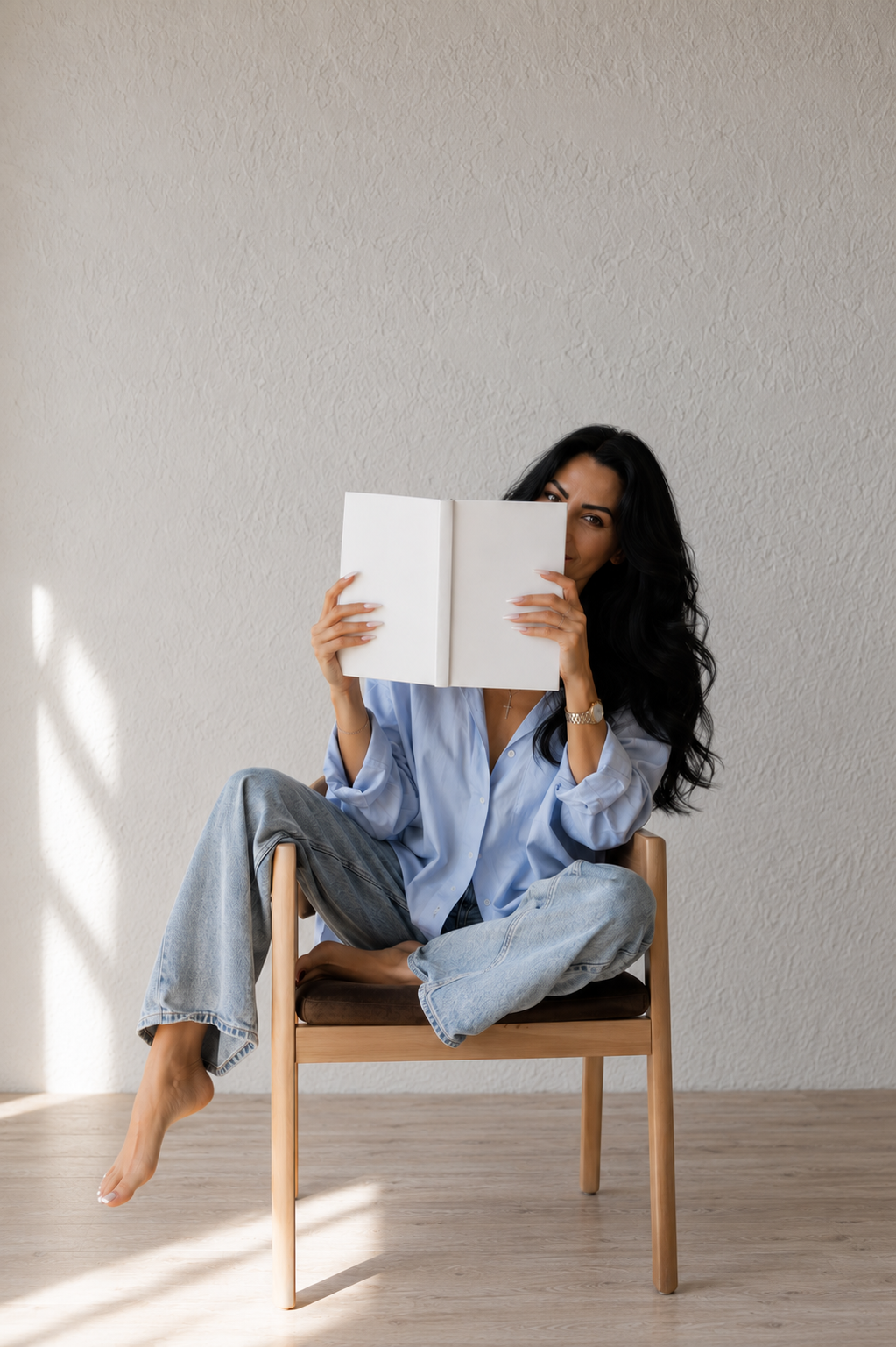 A woman with long dark hair wearing a blue shirt and ripped jeans sitting cross-legged on a wooden chair, holding and partially hiding her face behind an open white book, in a room with a plain textured wall.