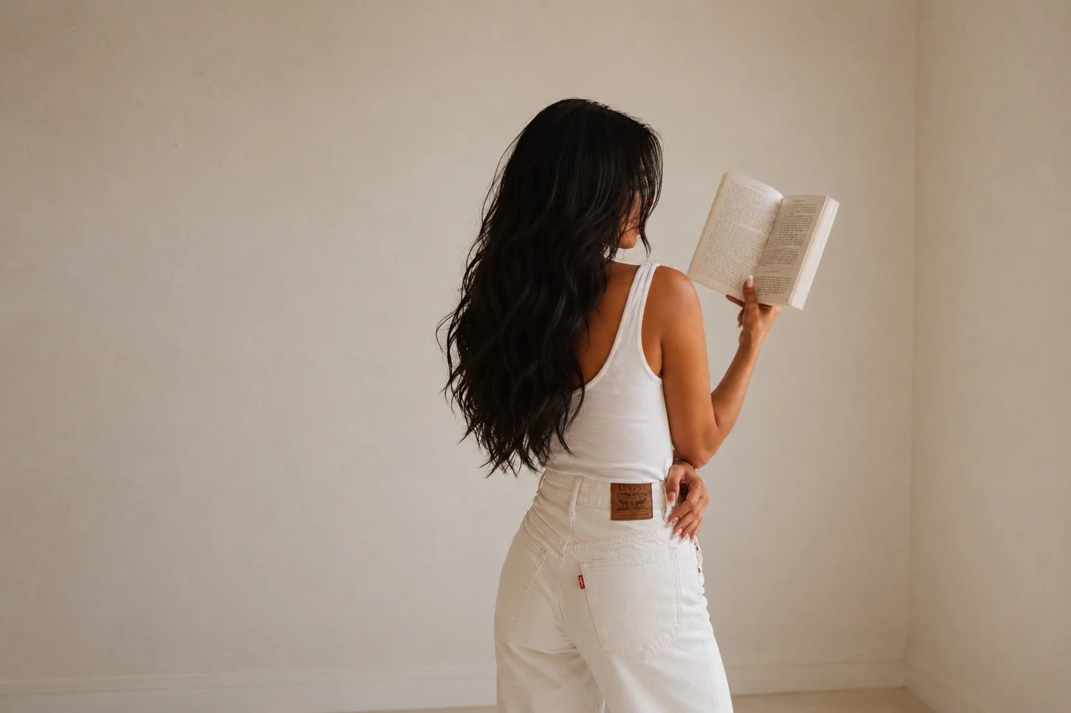 A woman with long dark wavy hair, wearing a white sleeveless top and white jeans, is standing in a minimalist room reading a book.