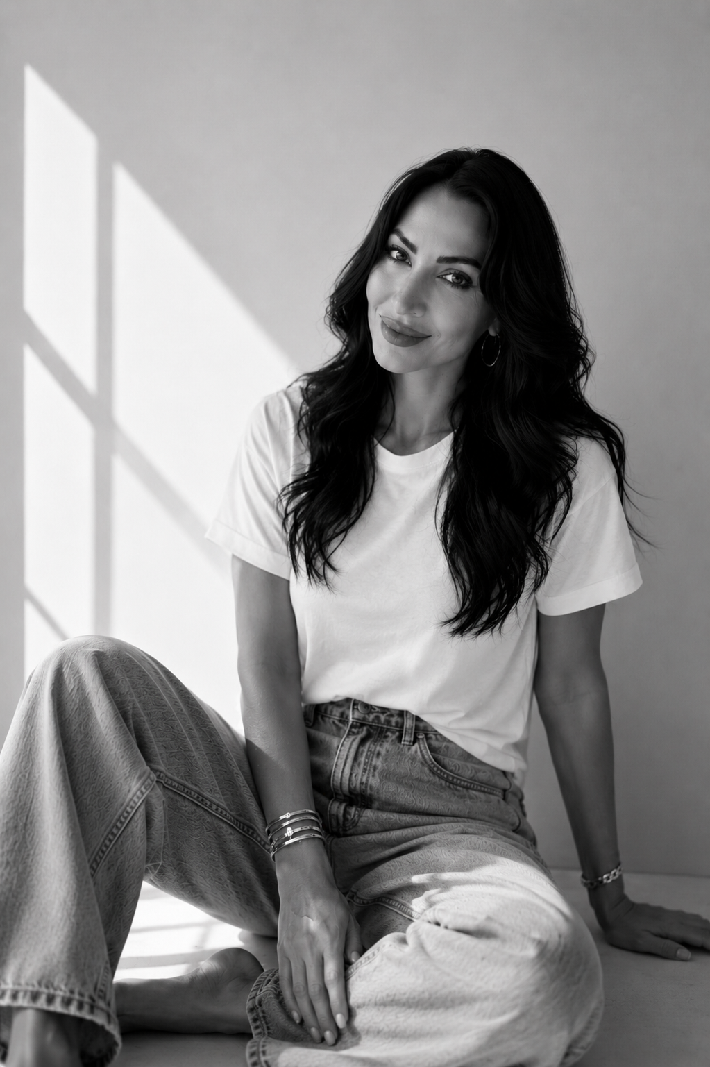 A woman with long wavy hair sitting on the floor, smiling at the camera, dressed in a white t-shirt and jeans, with sunlight casting shadows on the wall behind her.