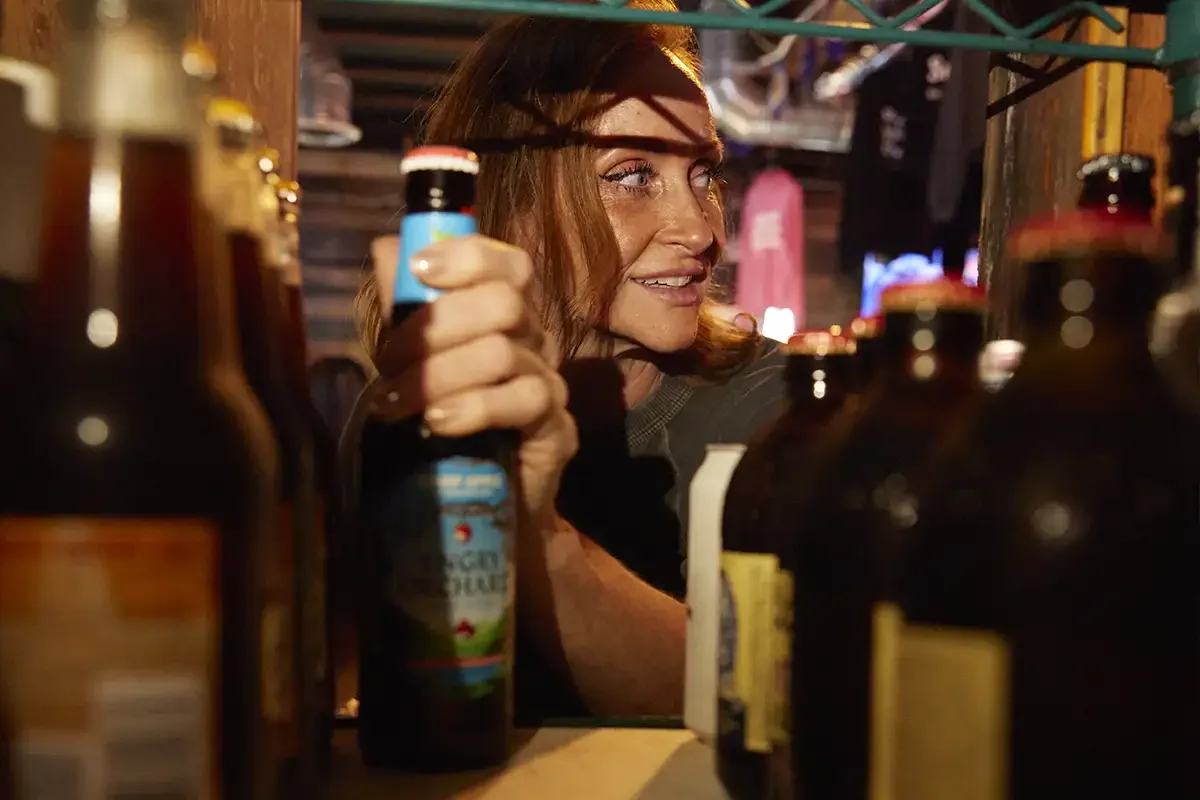 A woman at a bar or store shelf reaching for a bottle among other bottles, with warm lighting and a rustic wooden background.