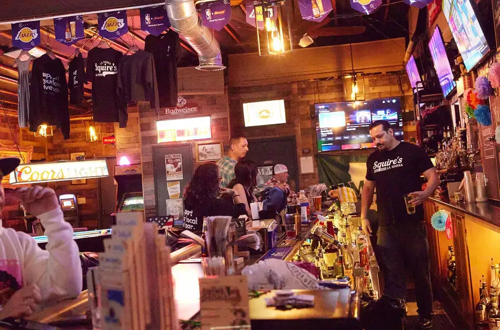 Inside a lively bar with wooden decor, patrons socializing, and a bartender serving drinks. T-shirts and banners hang from the ceiling, and multiple televisions display sports and entertainment.