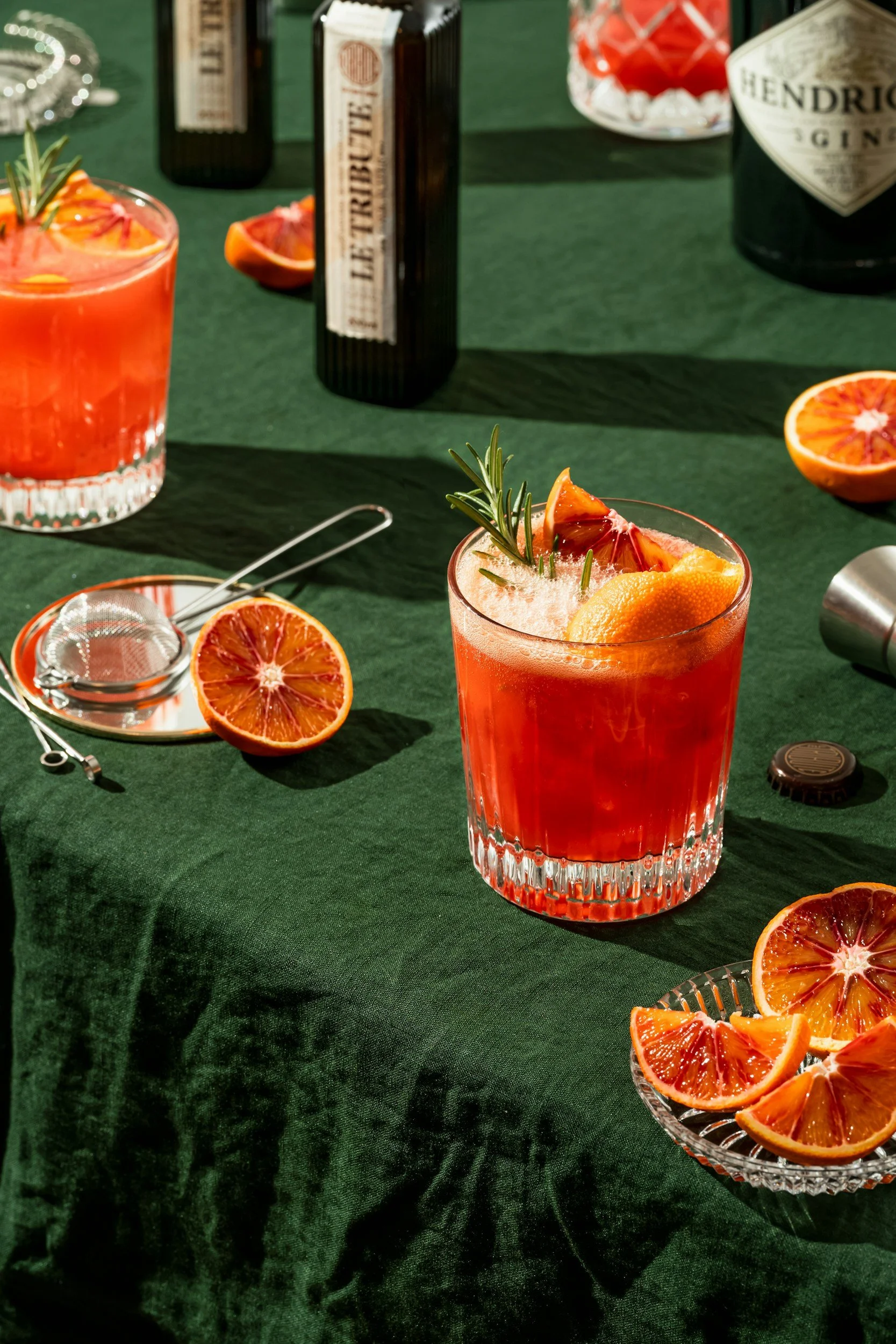 A glass of blood orange cocktail garnished with blood orange slices and rosemary sprig, on a dark green table with blood orange halves and slices, a citrus squeezer, and bottles of gin in the background.