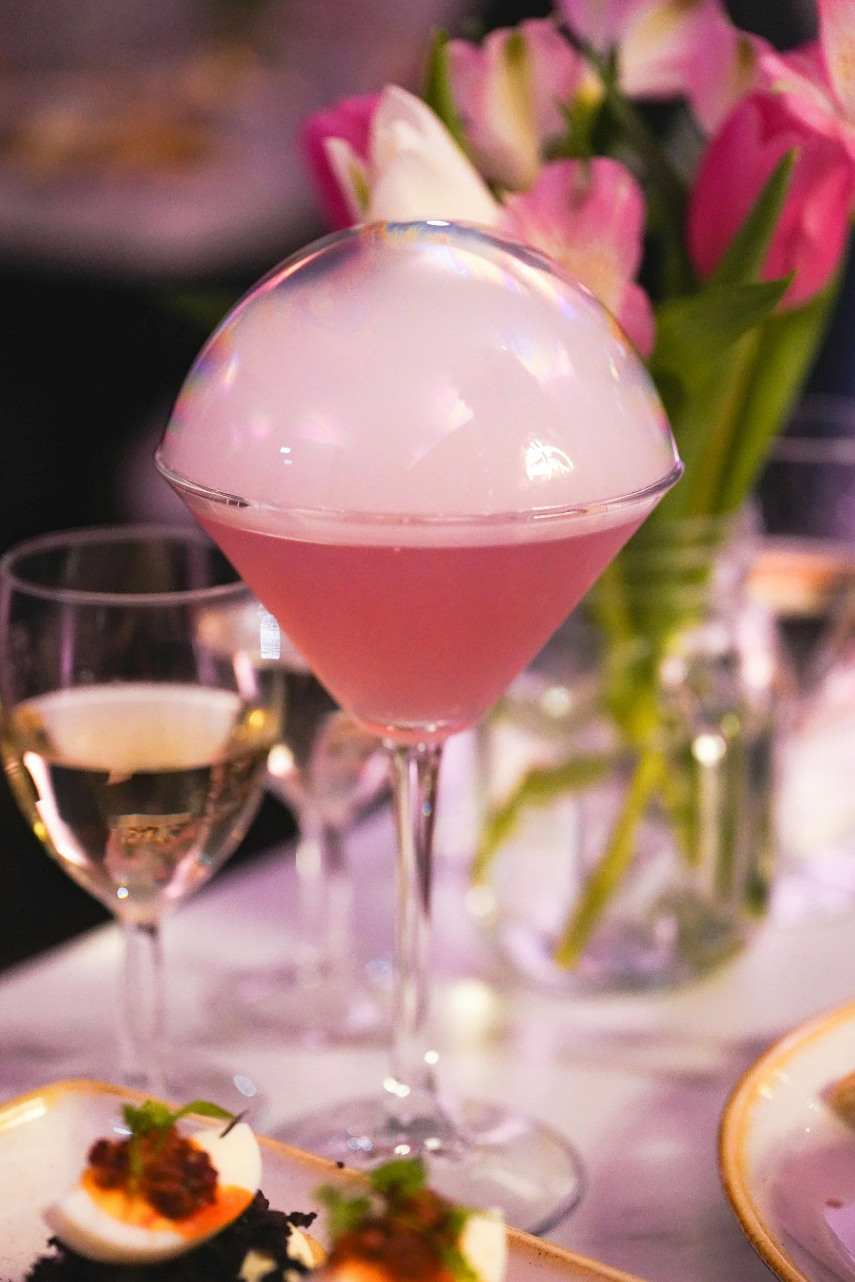 A cocktail glass filled with a pink drink topped with a large ice sphere. In the background, there are pink flowers and other glasses, suggesting a fancy event or celebration.