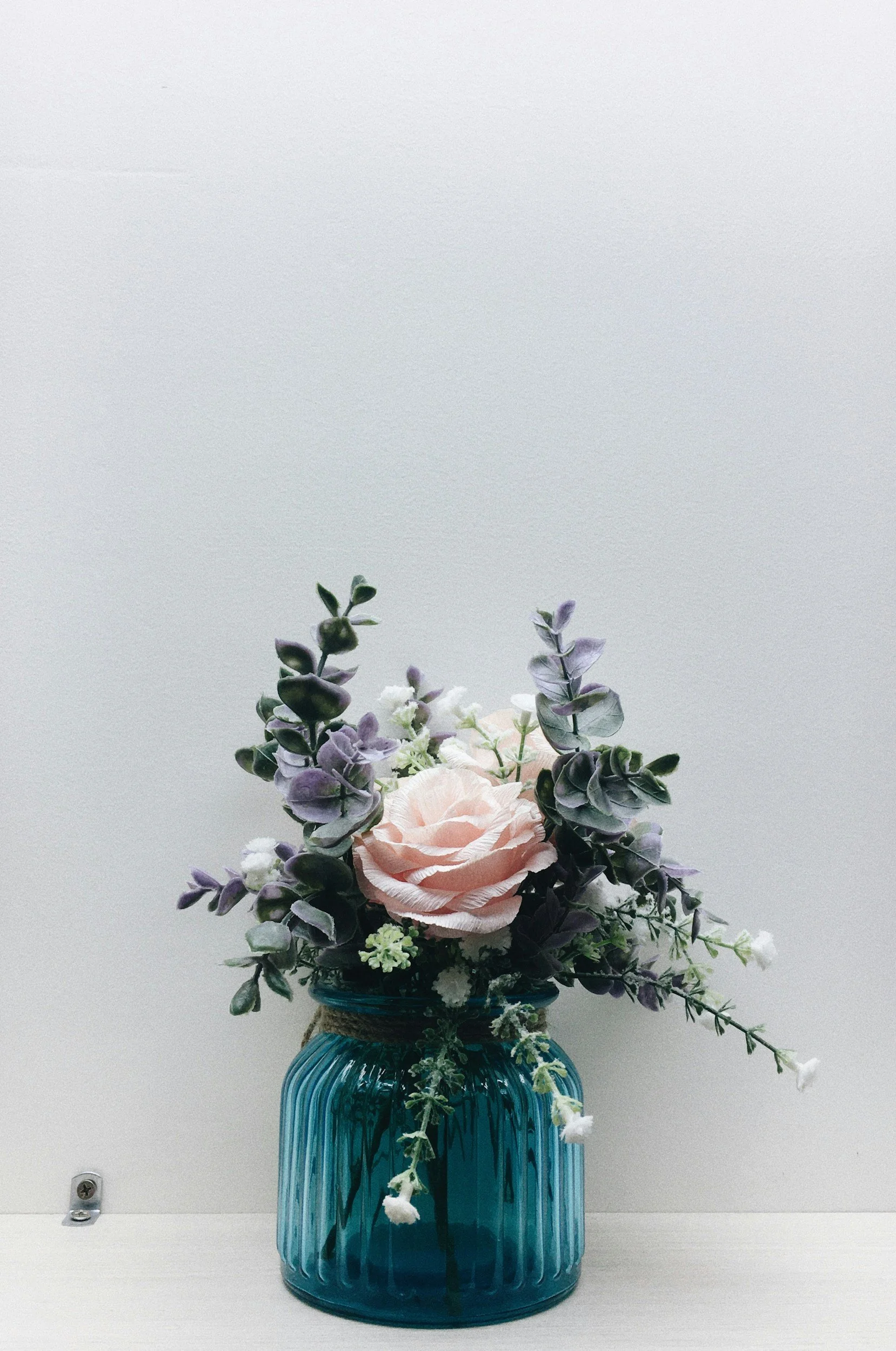 A floral arrangement in a blue glass vase containing pink roses, purple and green leaves, and white flowers, positioned on a white surface against a plain white background.