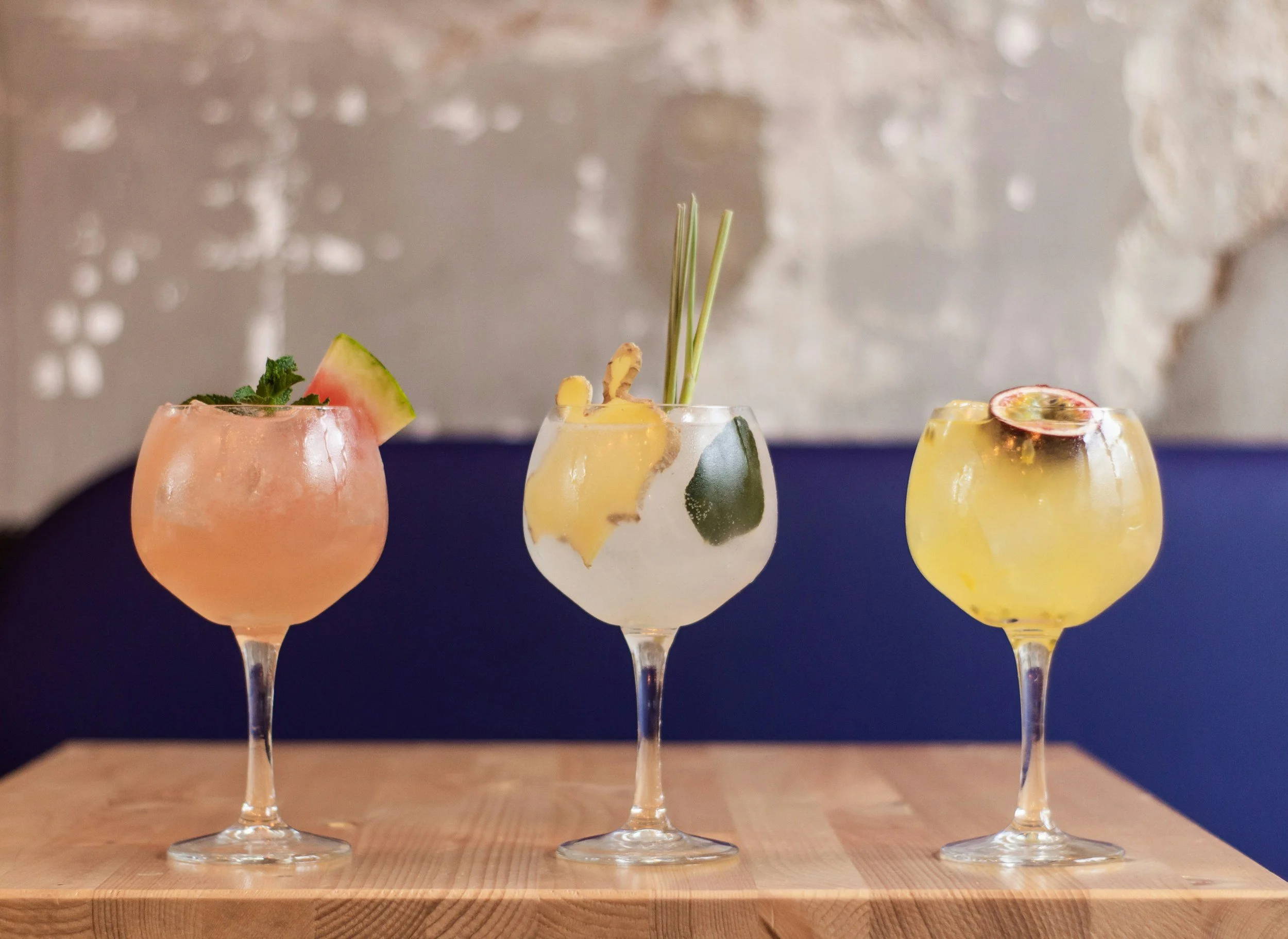 Three colorful cocktails in stemmed glasses on a wooden surface, with a blurred background.