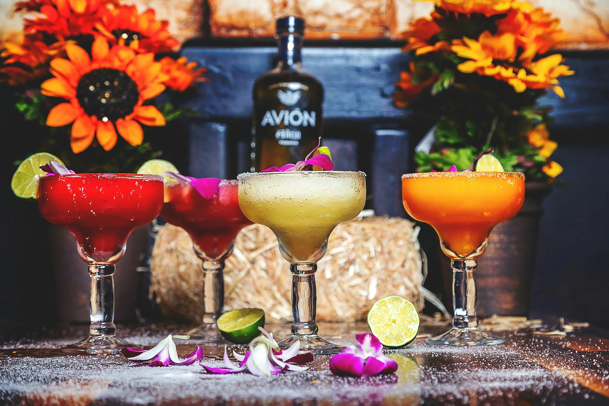 Four colorful cocktails on a bar counter with lime slices and flower petals, with bright orange flowers and a bottle of Avion tequila in the background.