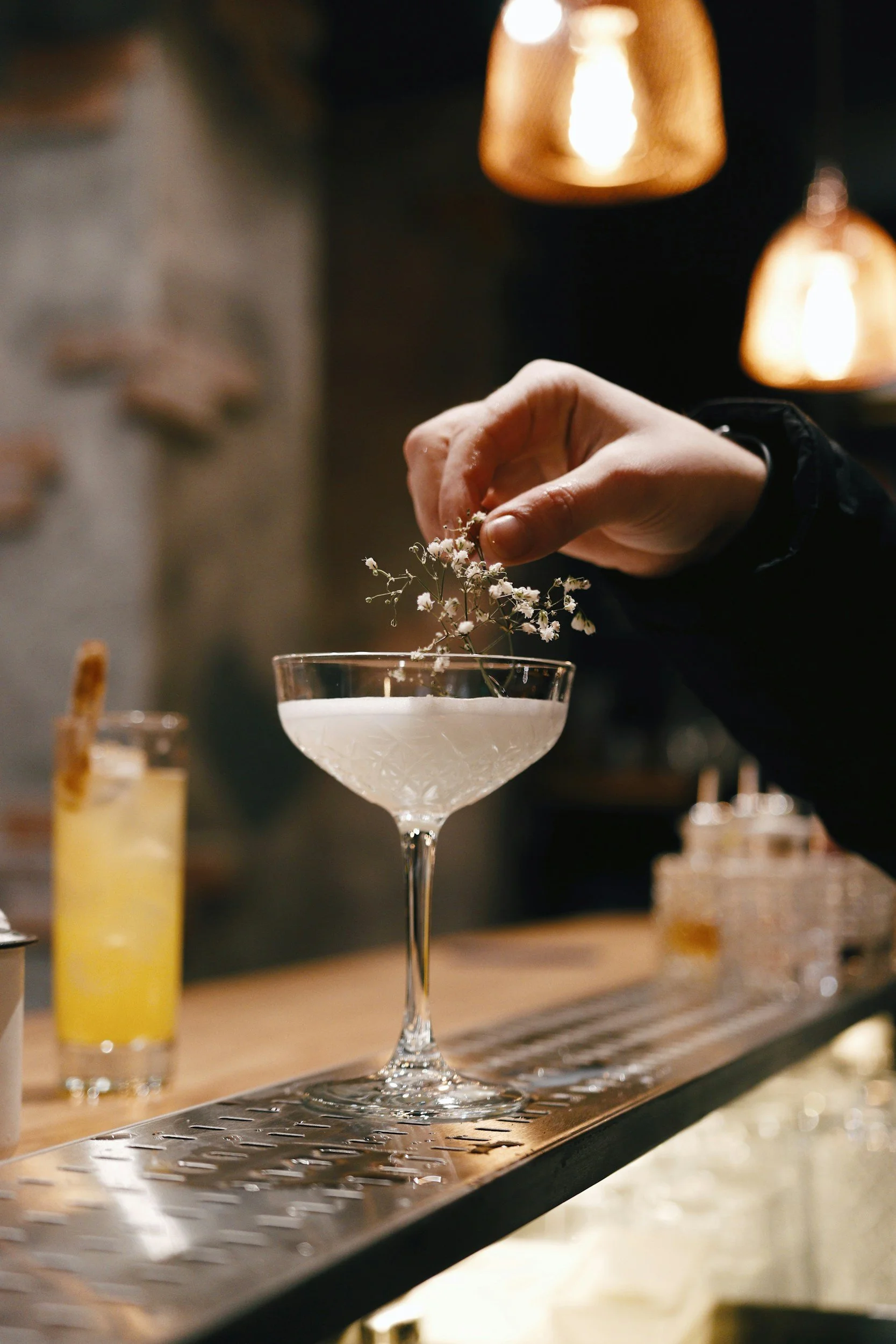 A person is placing a small white flower into a cocktail glass with a stem, on a bar counter. The background shows warm lighting and blurred drinks.