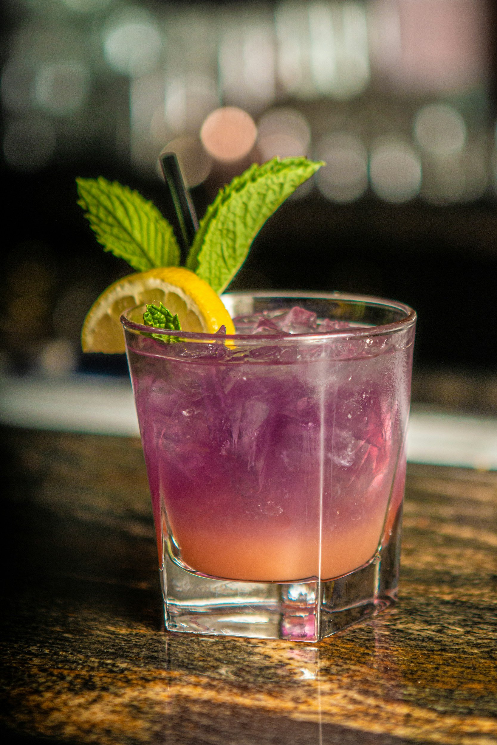 A pinkish-purple cocktail in a short glass with ice, garnished with lemon and mint, on a wooden surface.