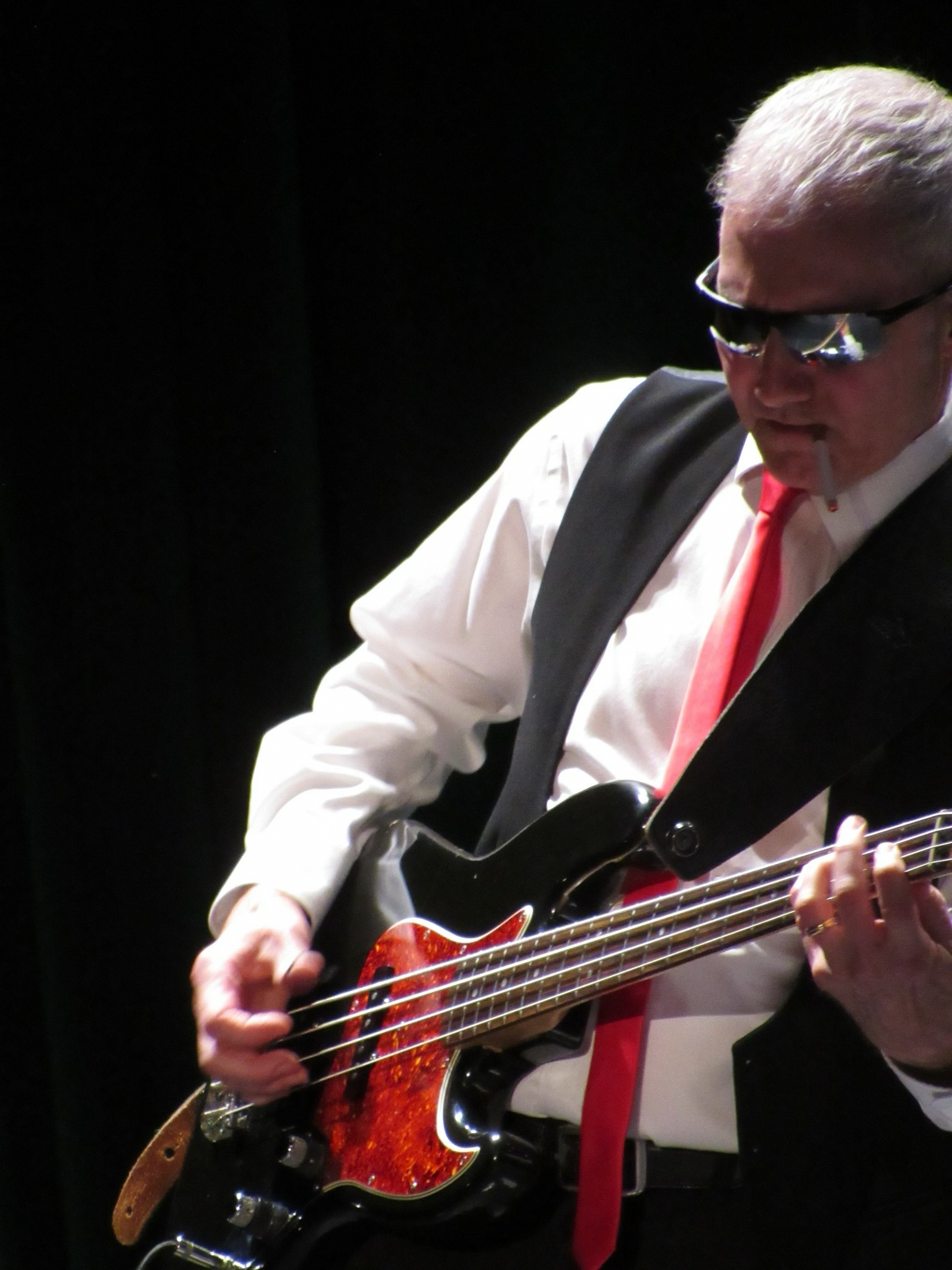 Man wearing sunglasses, a white shirt, black vest, and red tie standing in a music stage setting with equipment and colorful lights.