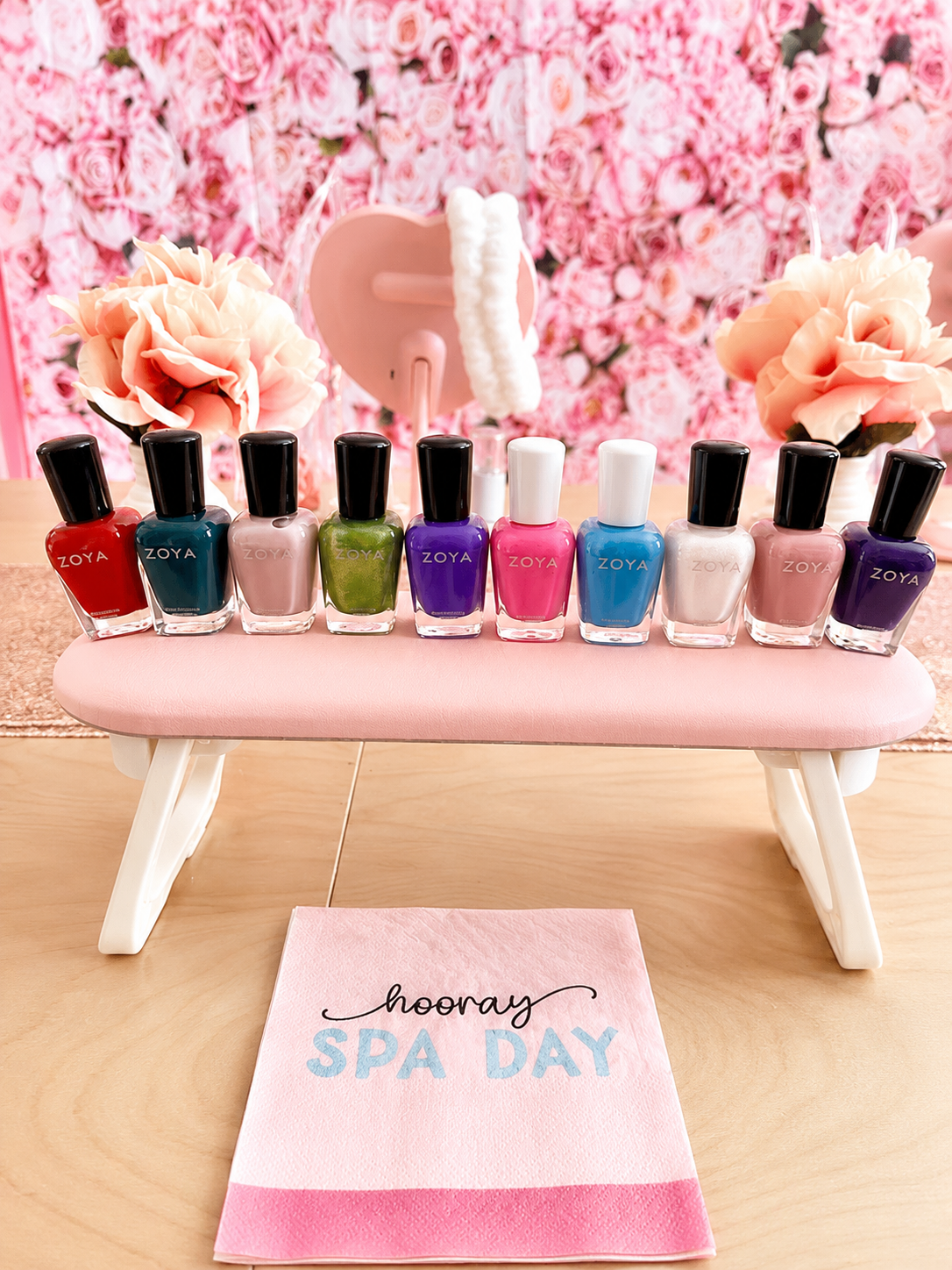 A display of ten colorful Zoya nail polish bottles on a pink mini bench, with pink roses and a floral pink wall in the background, and a pink card reading 'hooray SPA DAY' in front.
