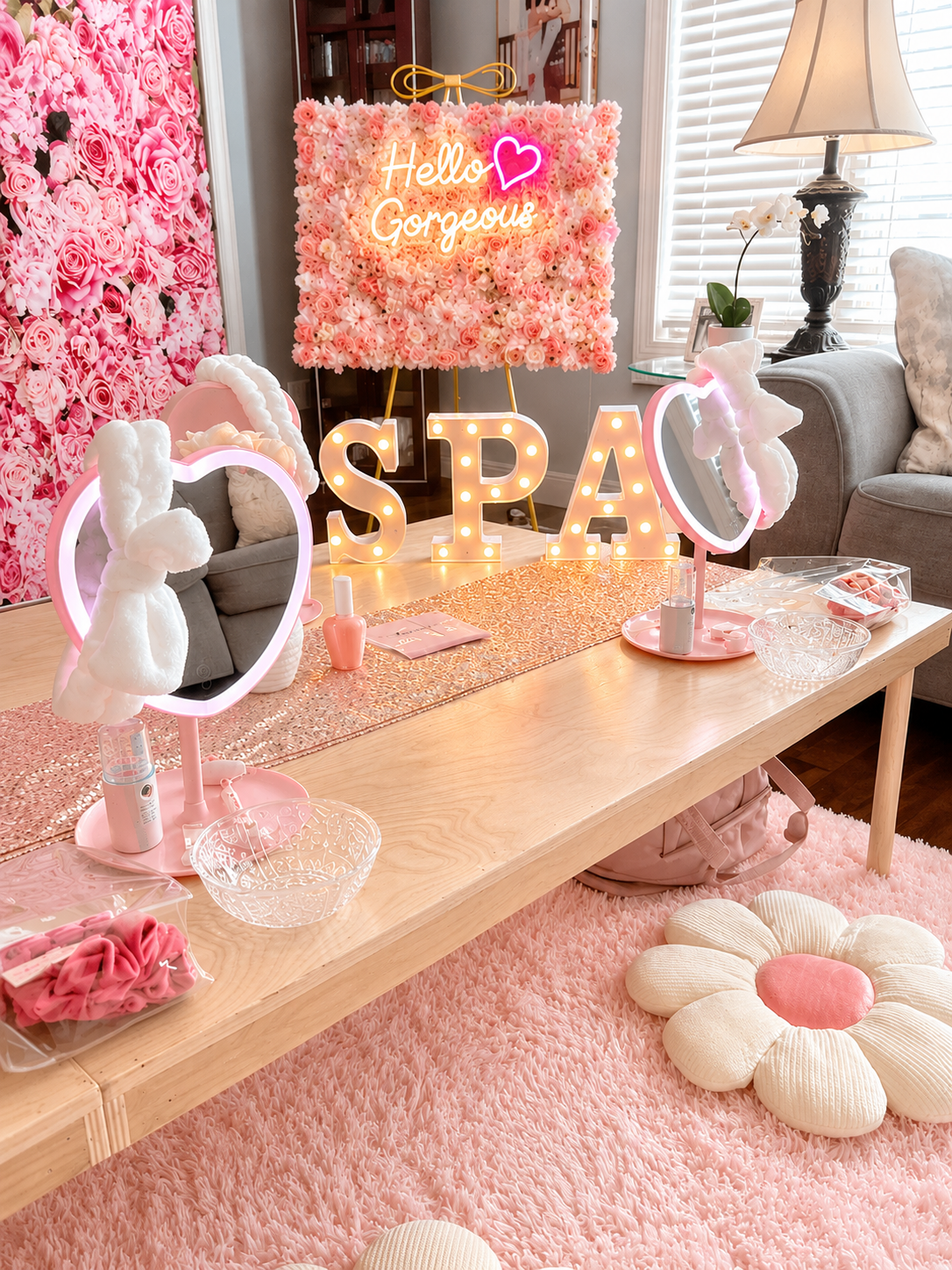 Decorated table with pink flowers, plush bunny-themed mirror lights, a glittery table runner, and a sign spelling 'SPA' with light bulbs. Pink roses wall, neon sign reading 'Hello Gorgeous' with a heart, and a cozy pink flower-shaped floor cushion.