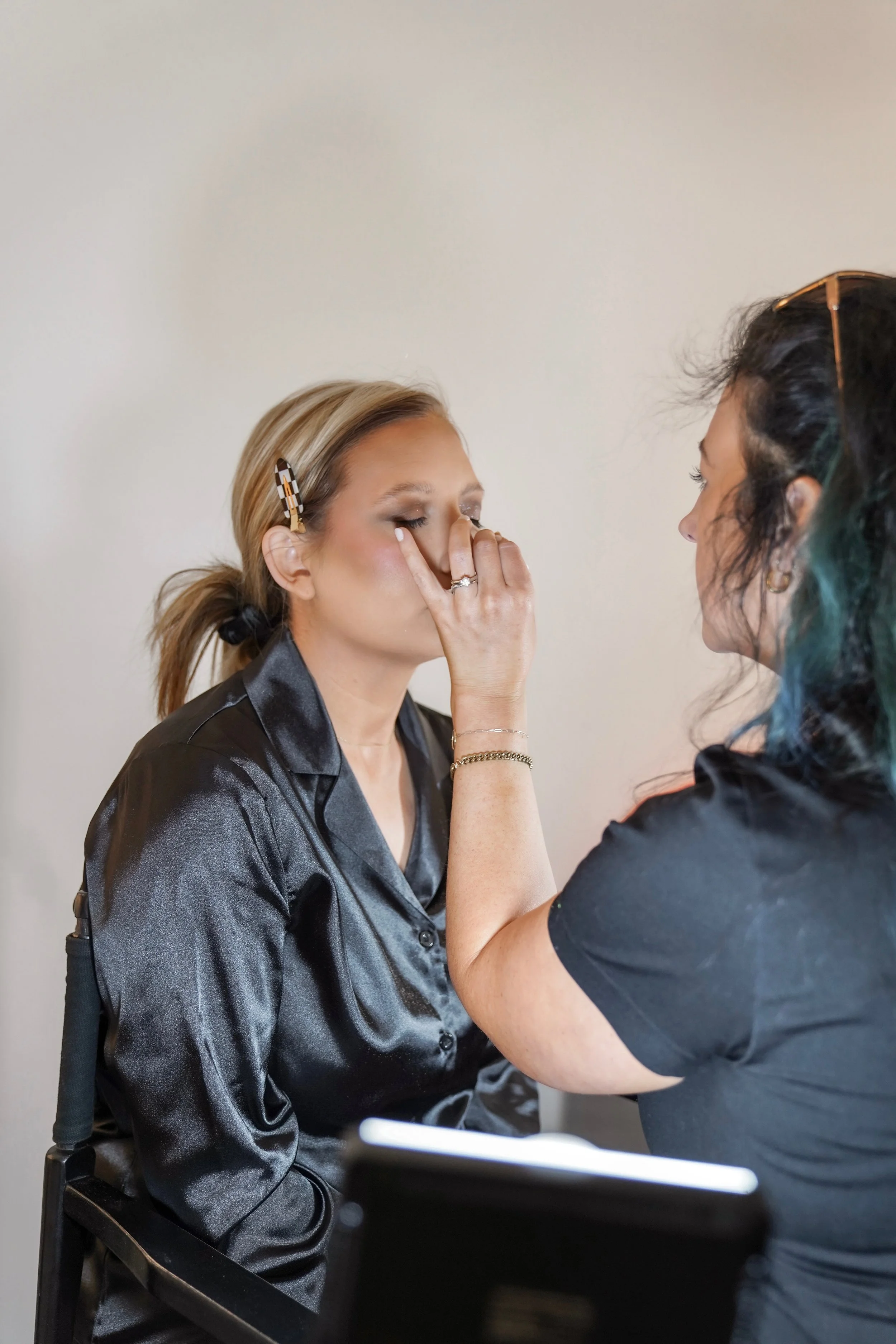 A woman sitting in a chair having makeup applied by a makeup artist. The woman has blonde hair with a hair clip and is wearing a black satin button-up shirt. The makeup artist has dark hair with a hint of teal. They are both focused on the makeup application.